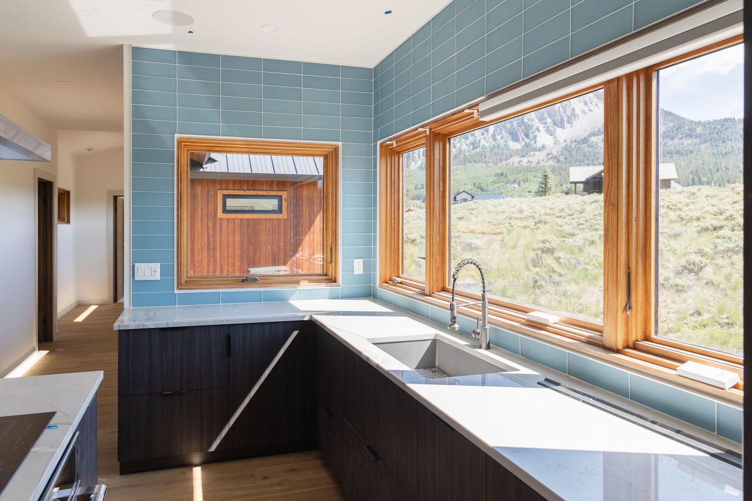 Modern kitchen with dark cabinets, stainless steel sink, blue tile backsplash, and wood-framed windows overlooking a scenic mountain view.