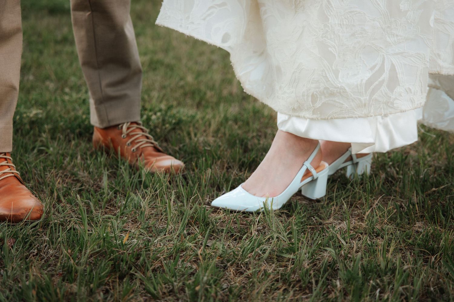 Close-up of bride in white dress wearing light blue heels and groom in tan pants with brown leather shoes on grass.