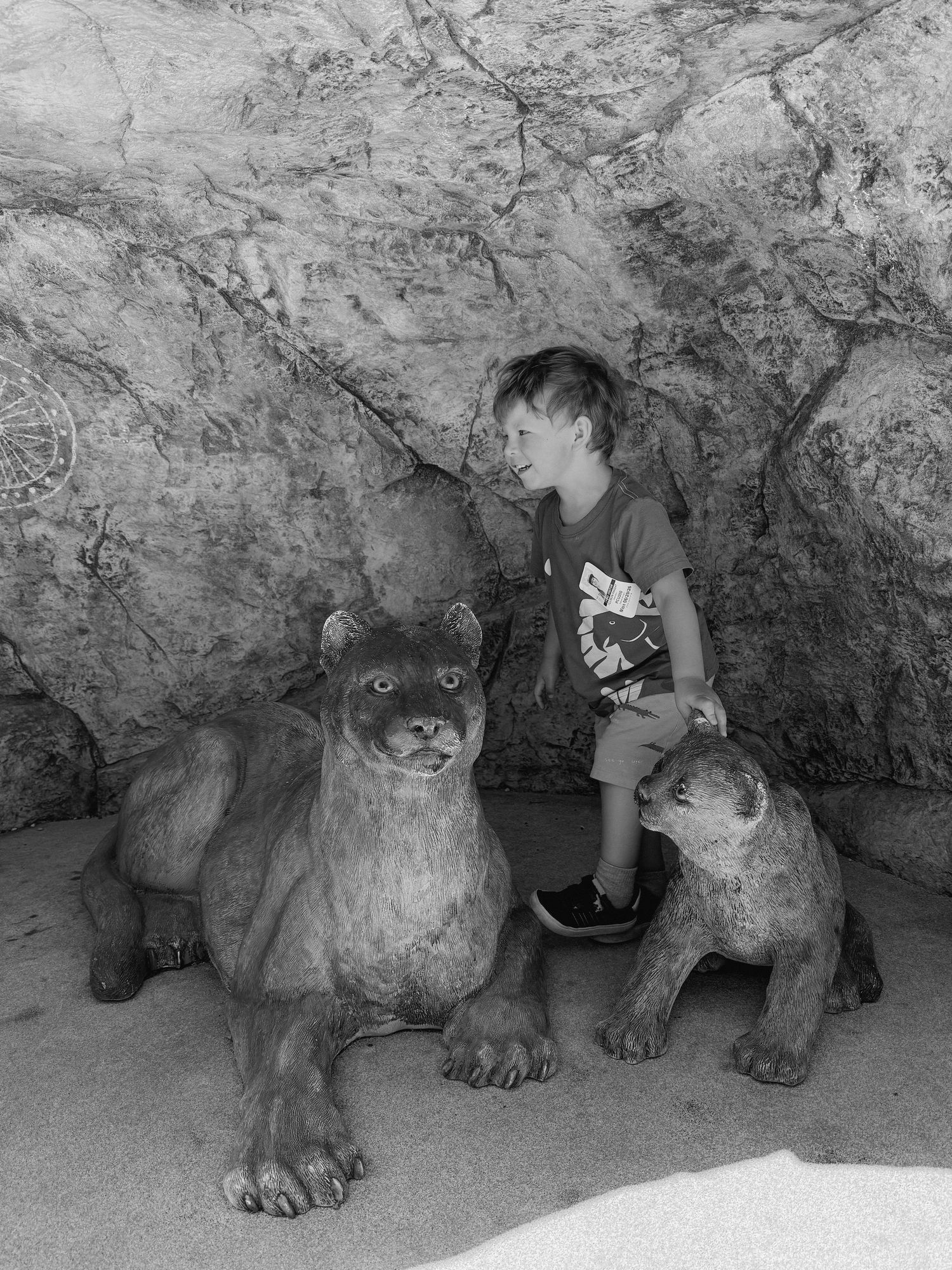 Boy sitting on a large stone mountain lion sculpture with a smaller cub statue in a rocky cave setting.