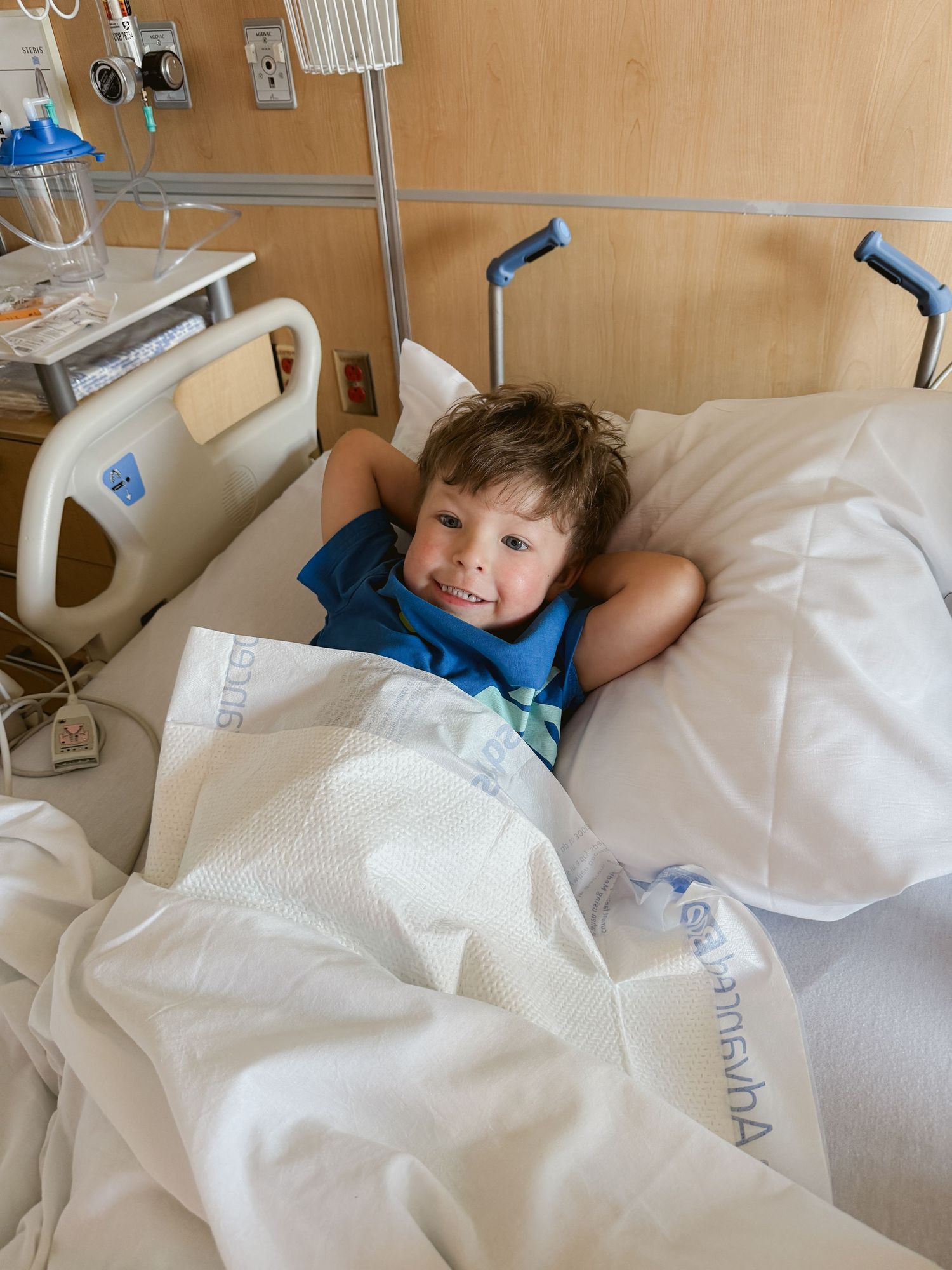 Young smiling boy resting in a hospital bed, covered with white blankets, wearing a blue shirt.