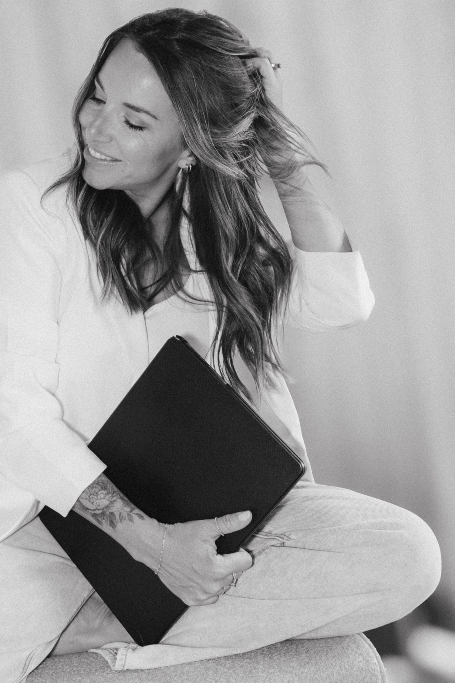 Smiling woman with tattoos holding a black folder while sitting cross-legged.