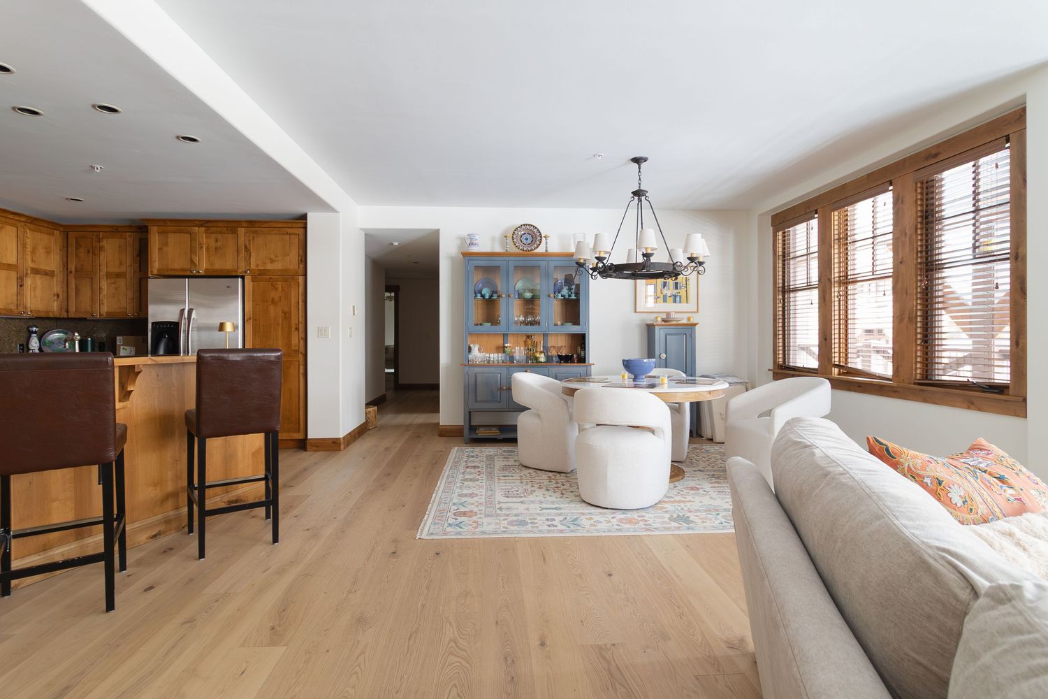 Open-concept living room with light hardwood floors, beige sofa, wooden kitchen cabinets, and a dining area with chandelier.
