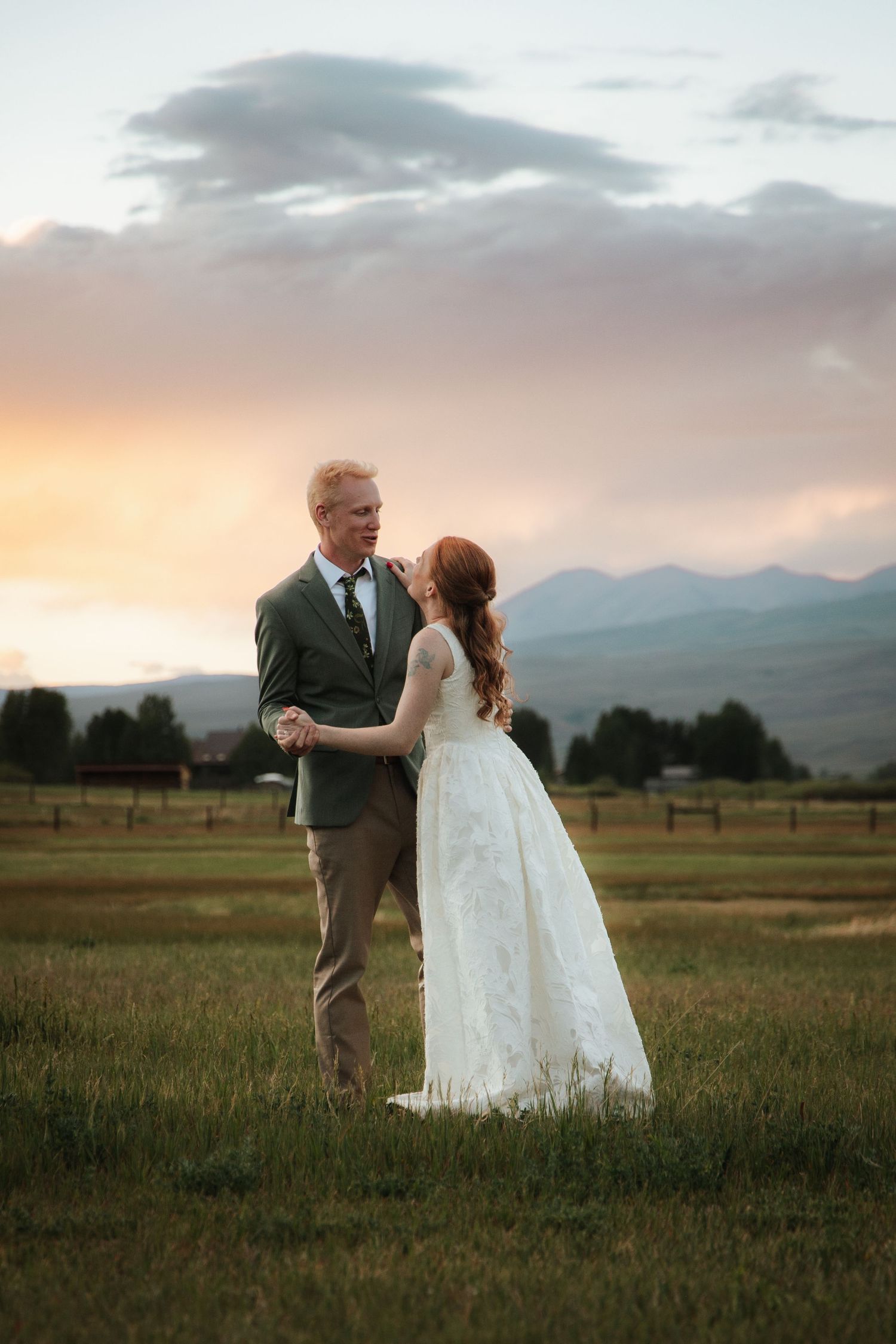 Bride and groom dancing in a green field at sunset with mountain backdrop on their wedding day.