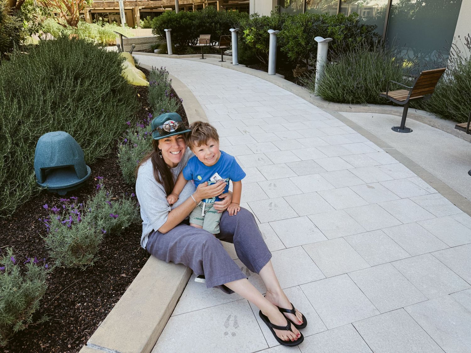 Woman and young boy sitting together on a garden pathway surrounded by lavender plants and greenery.