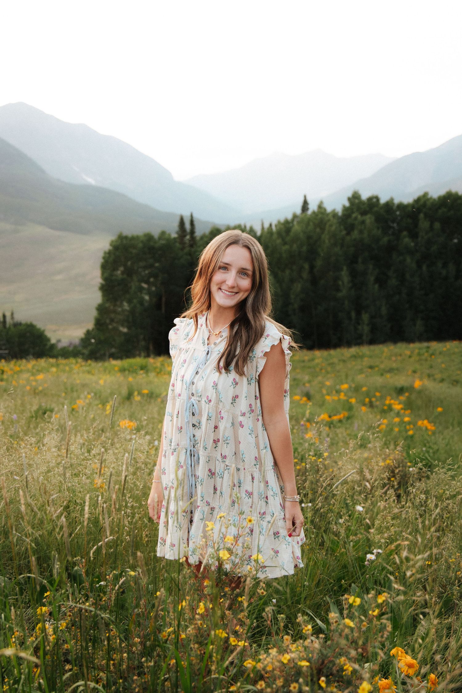 Young woman in floral dress smiling in a wildflower meadow with mountains and evergreen trees in the background.