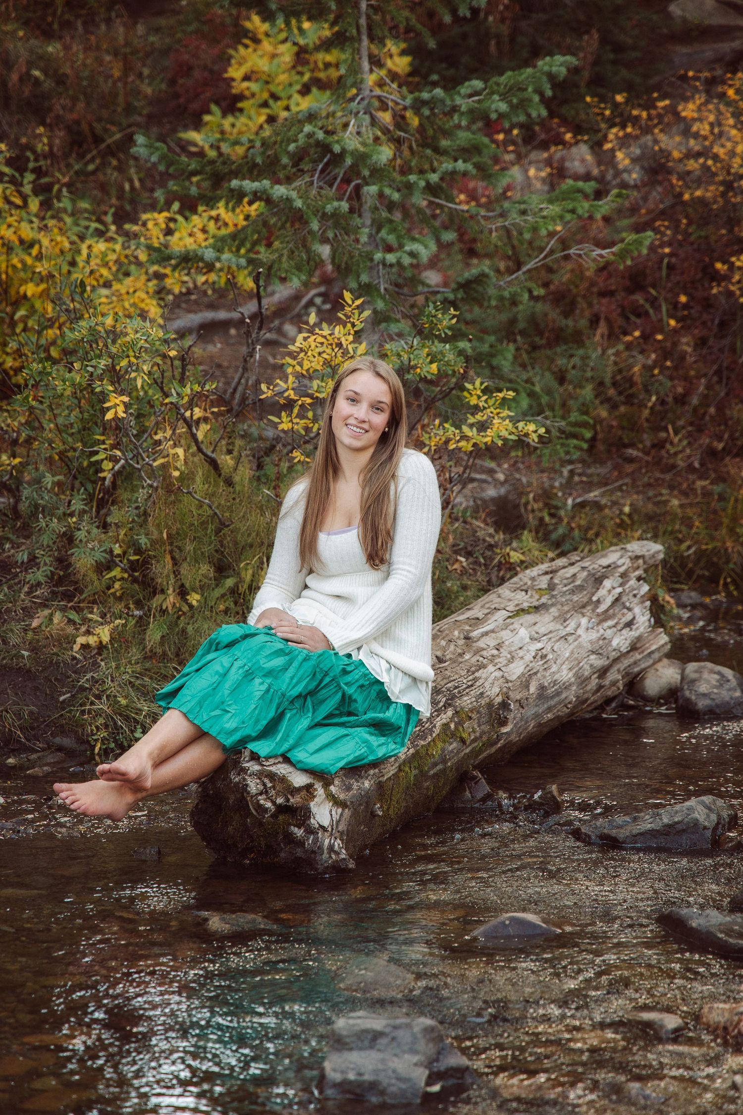 Person in white top and green skirt poses by stream with autumn foliage in background.