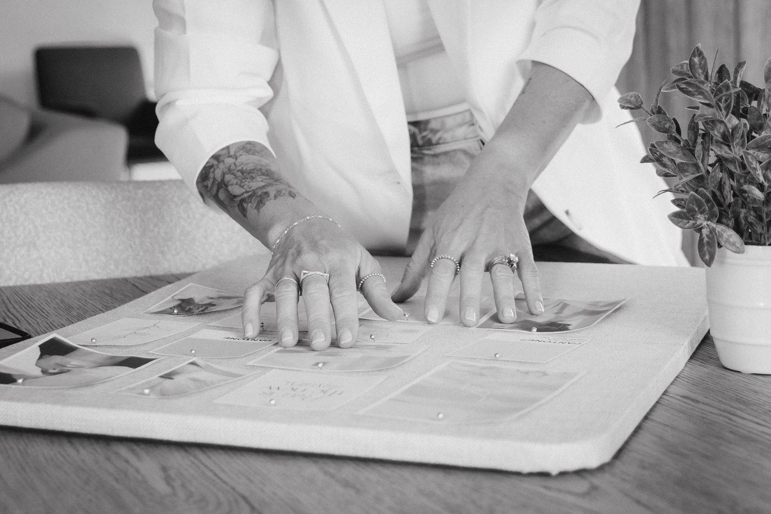 Person with tattooed hands reading a newspaper on a wooden table beside a small green plant.