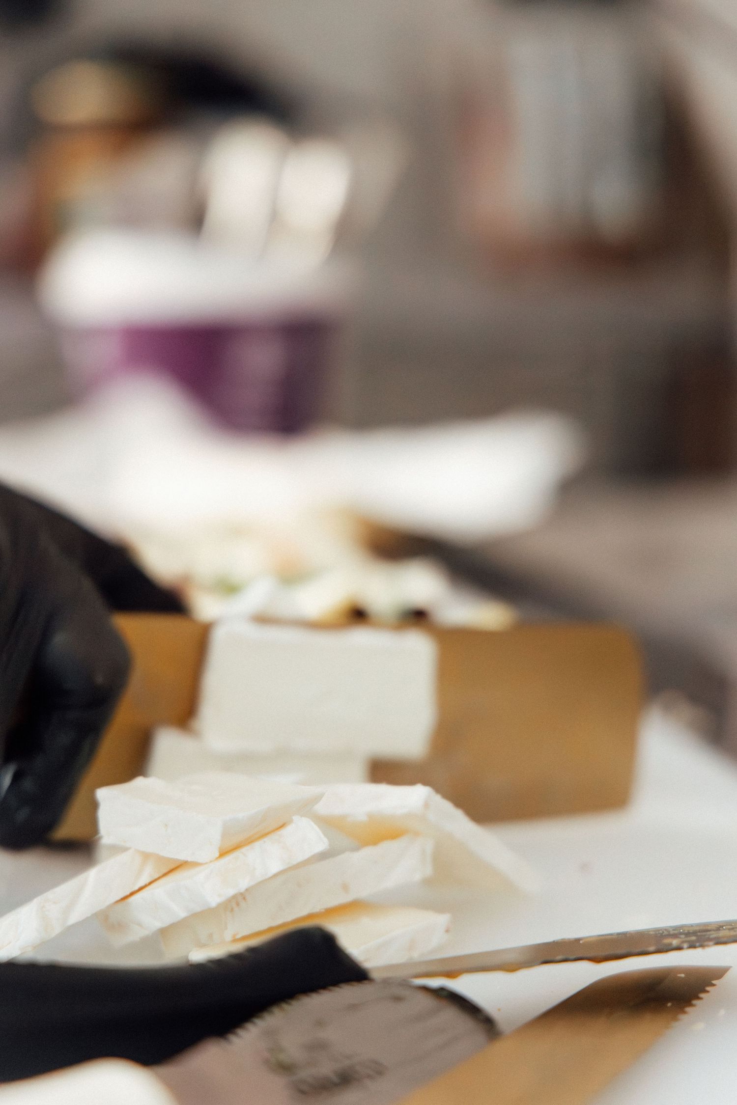 Gloved hands slicing fresh brie cheese on a wooden cutting board with a serrated knife.