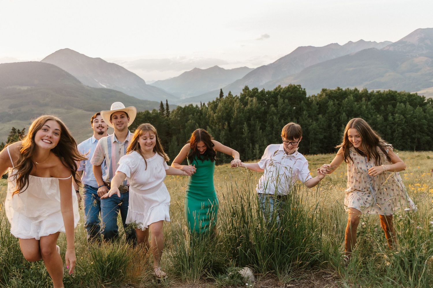 A group runs together through a mountain meadow at sunset wearing light summer clothing.