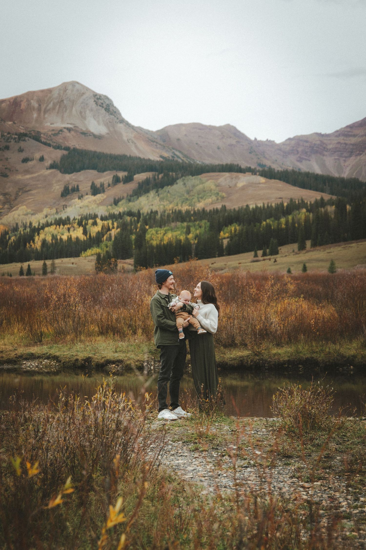 A young family holds their baby near a mountain lake surrounded by autumn foliage in Colorado.