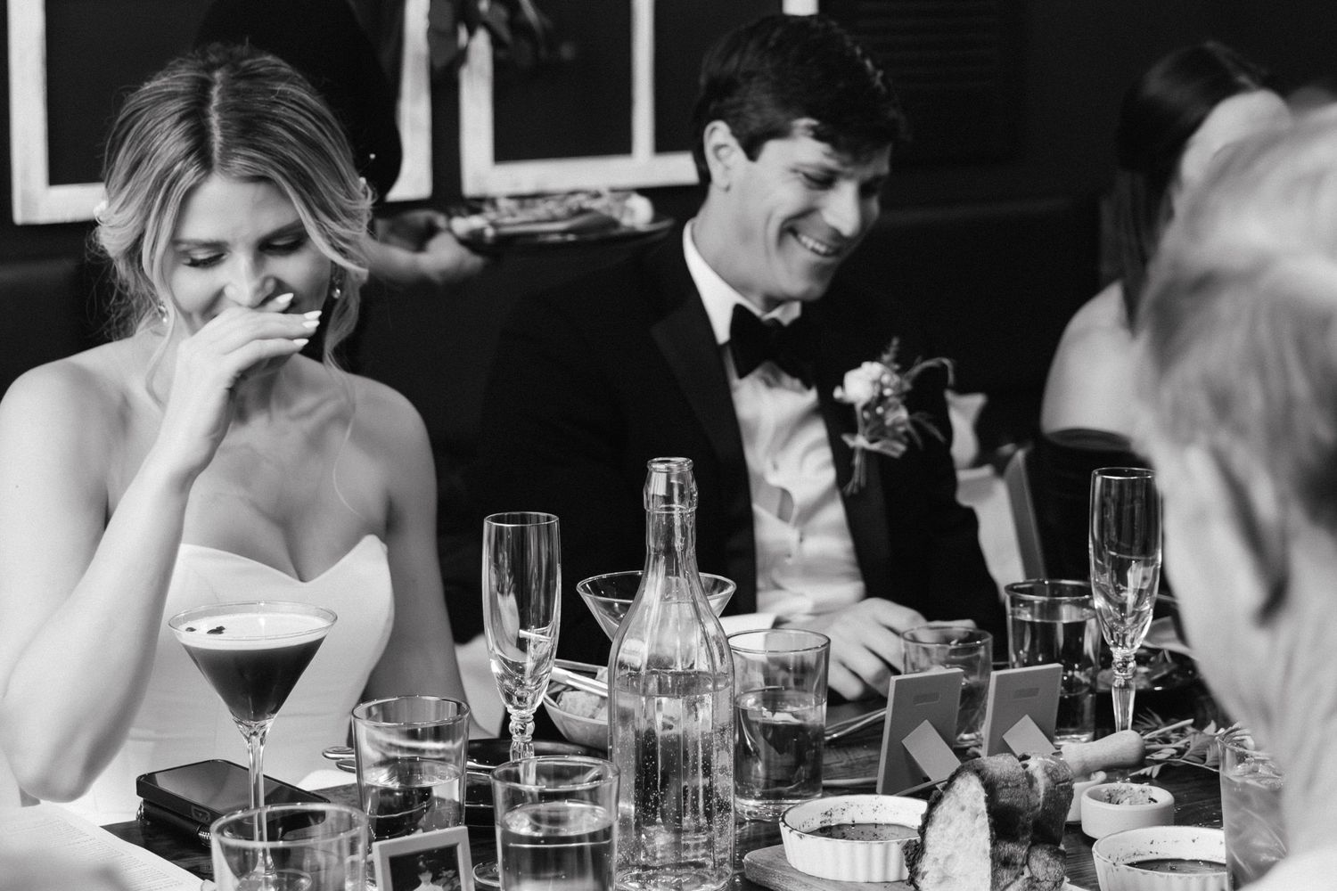 A bride and groom laugh joyfully at their wedding reception dinner table in black and white.