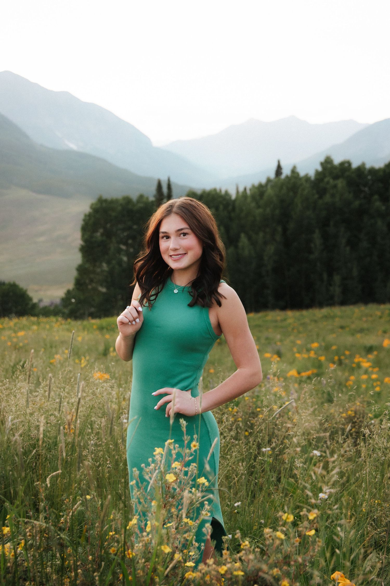 Young woman in green dress posing in mountain wildflower meadow with evergreen trees and misty peaks behind her.