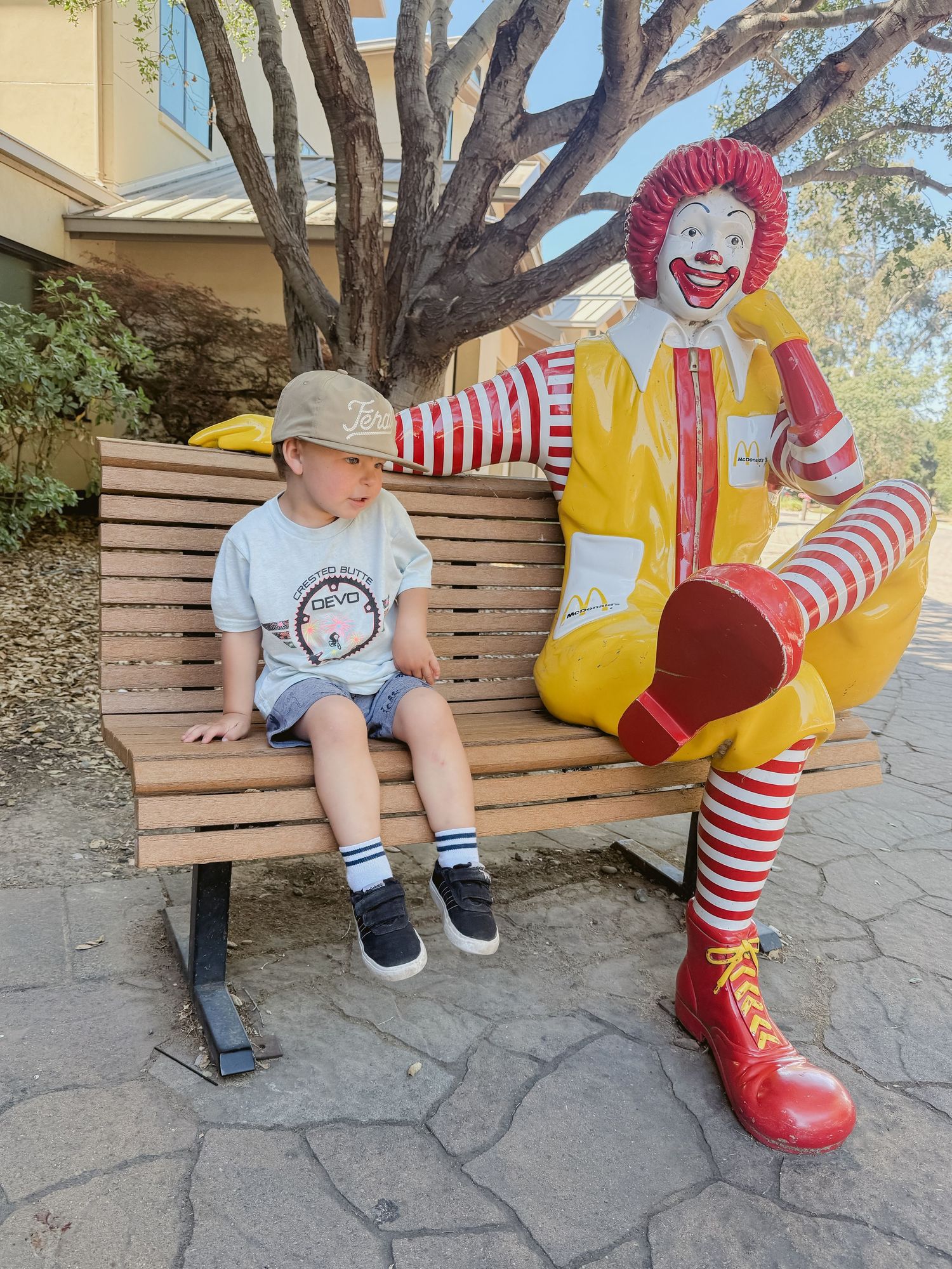 Young boy sitting on a bench beside a Ronald McDonald statue outside a McDonald's restaurant.