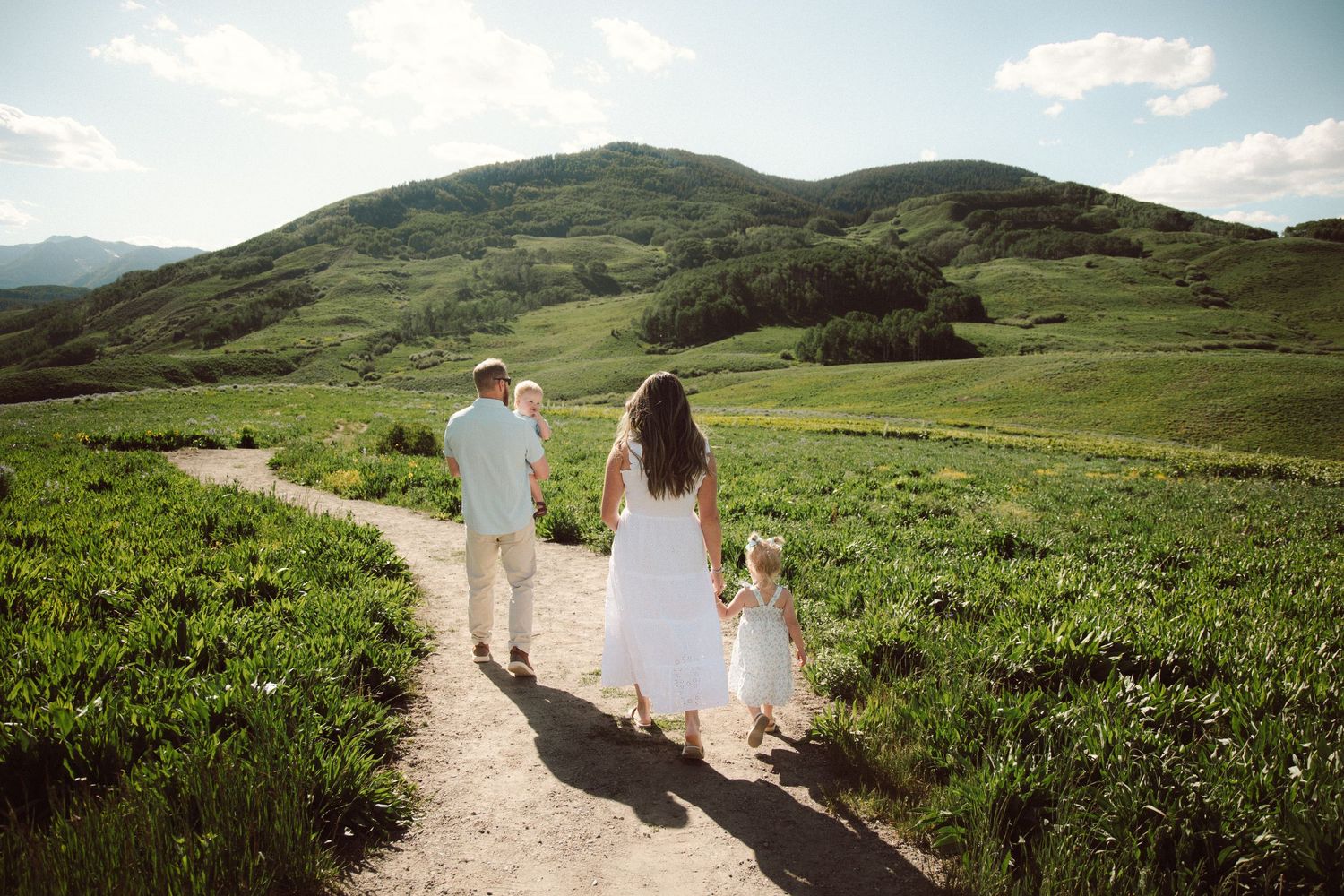 Family of three in white walking a green meadow path with rolling hills under a bright sky.