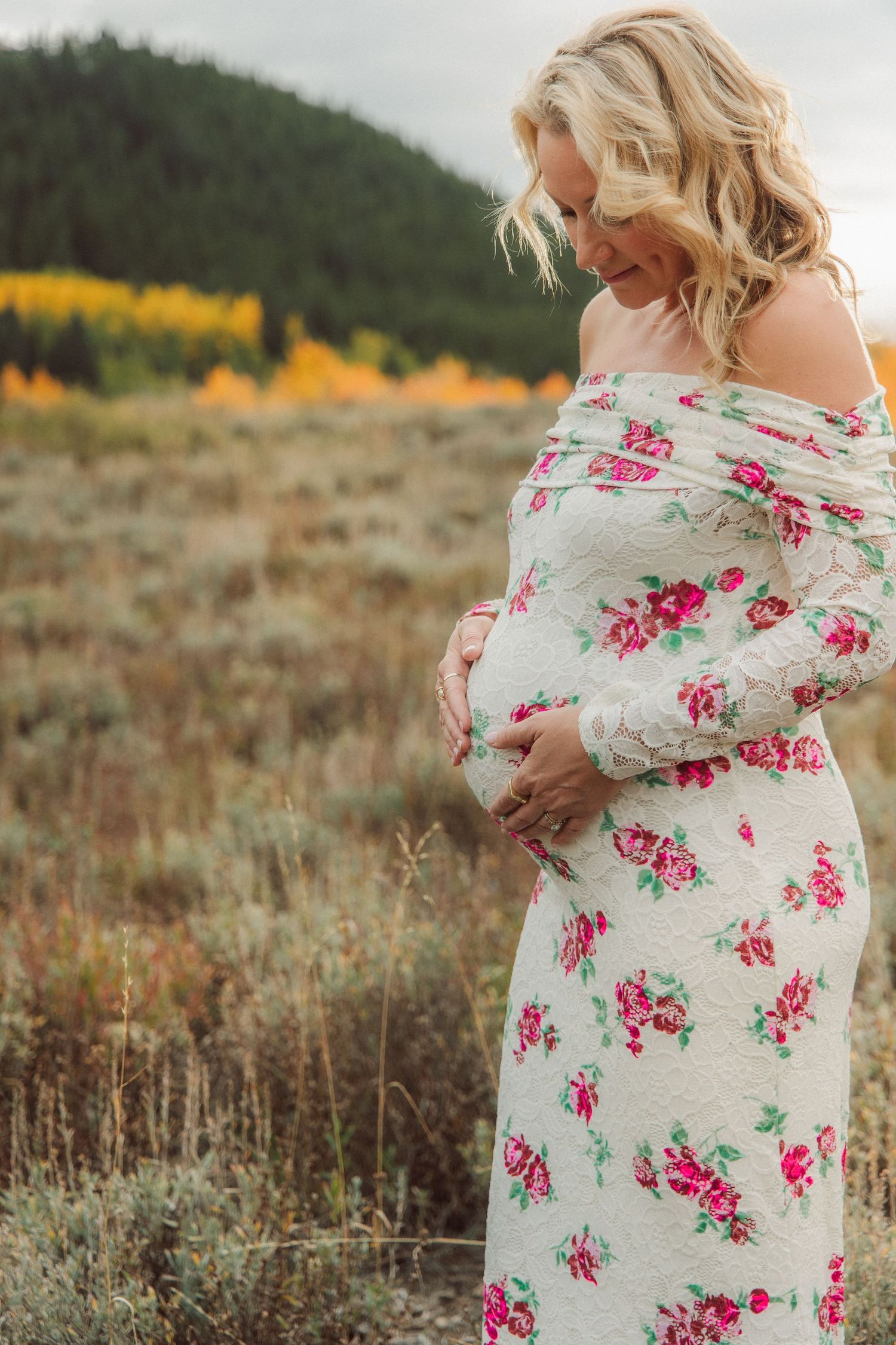 A floral maternity dress is showcased against a rustic autumn landscape with mountains in the background.