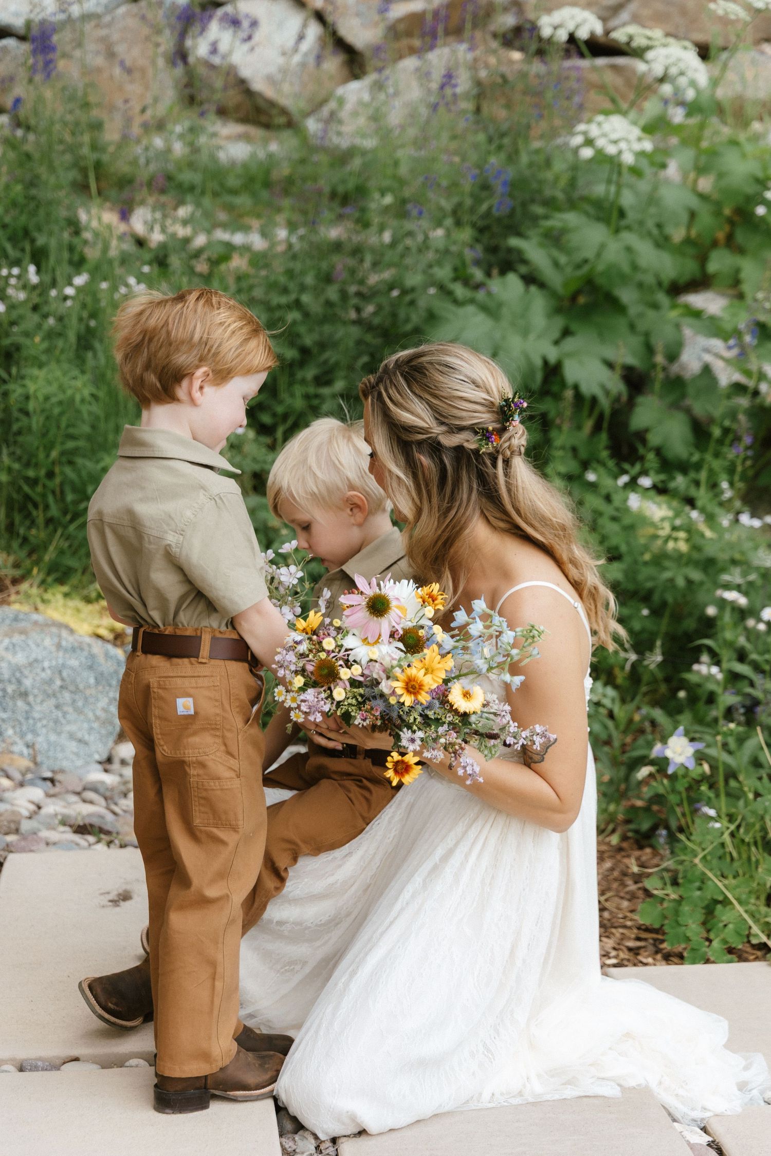 Bride in white gown kneels with two young boys smelling a colorful wildflower bouquet in a lush garden setting.