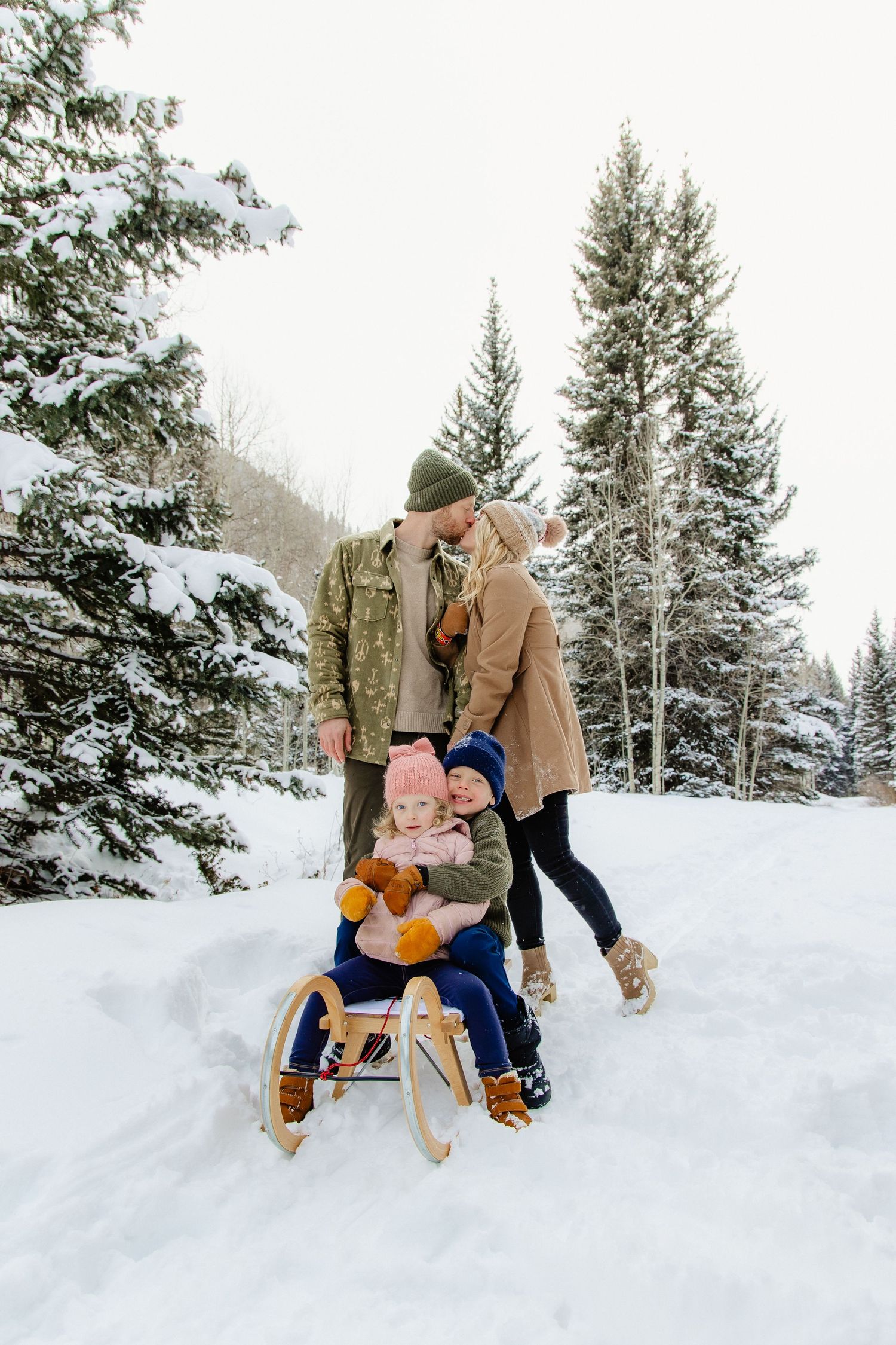 A family enjoys winter sledding together on a snow-covered path through evergreen forest.