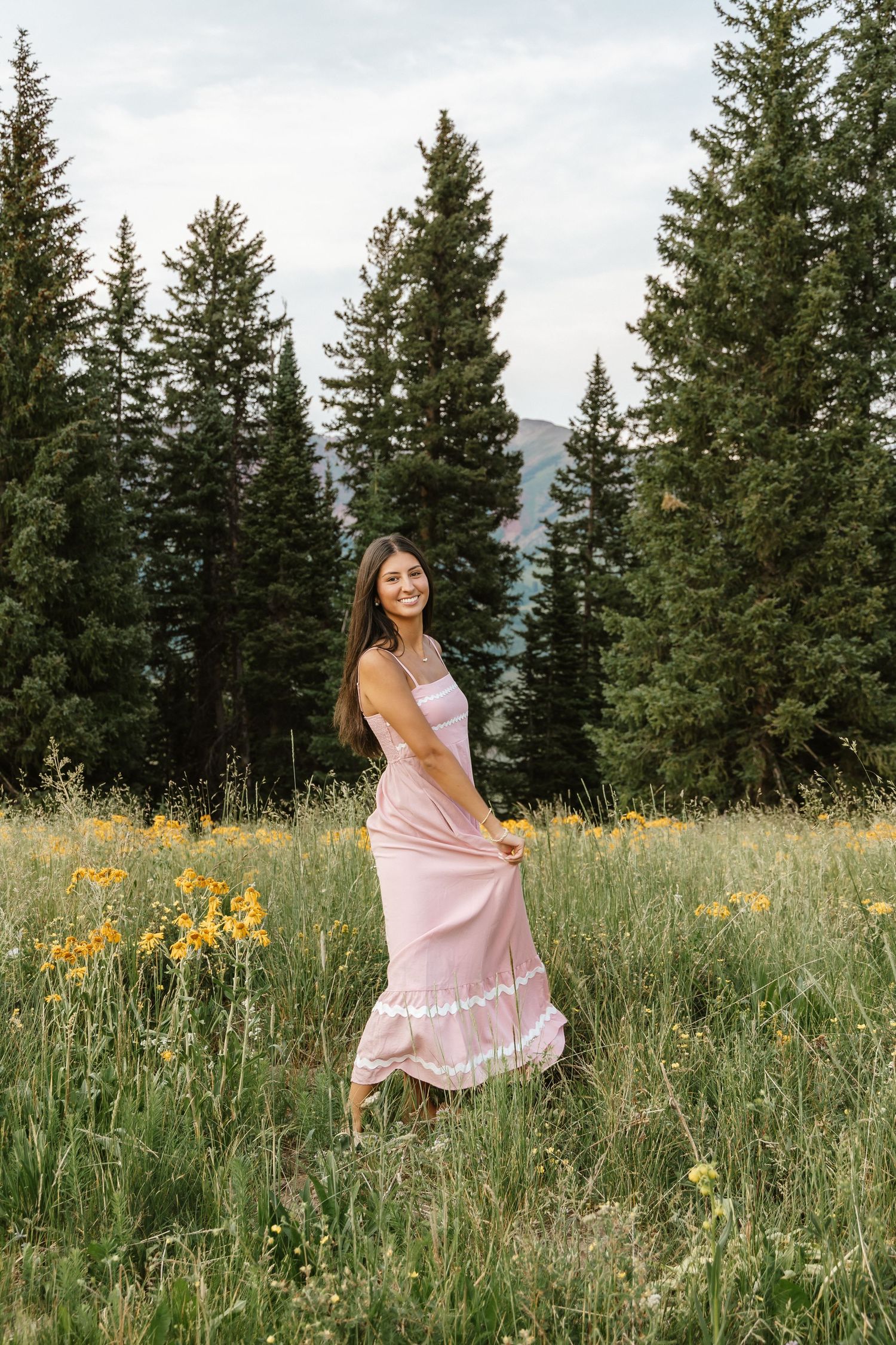 A woman in a pink dress twirls in a mountain meadow surrounded by evergreen trees and wildflowers.