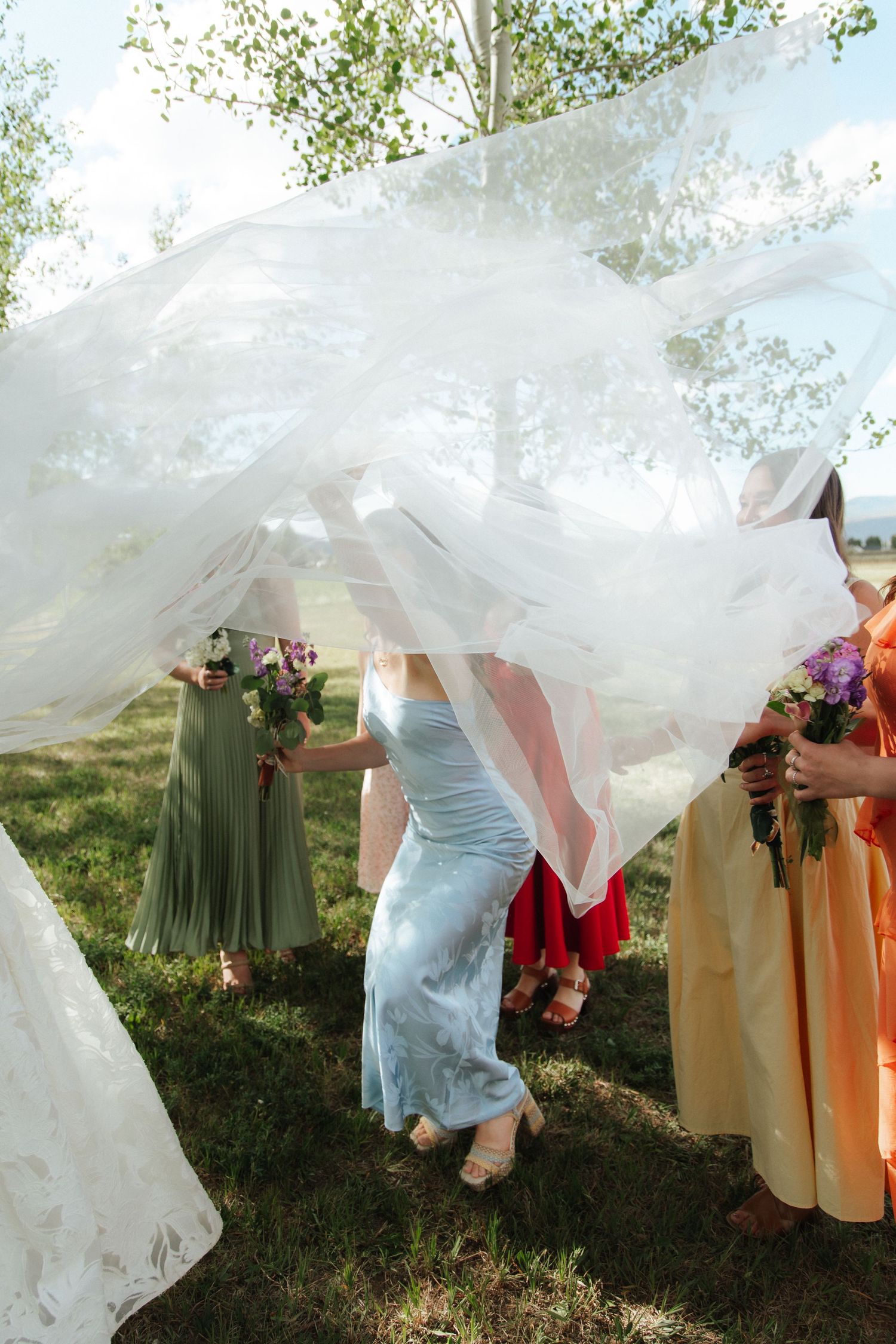 Bridesmaids in colorful dresses hold bouquets as a white wedding veil billows in the wind outdoors.