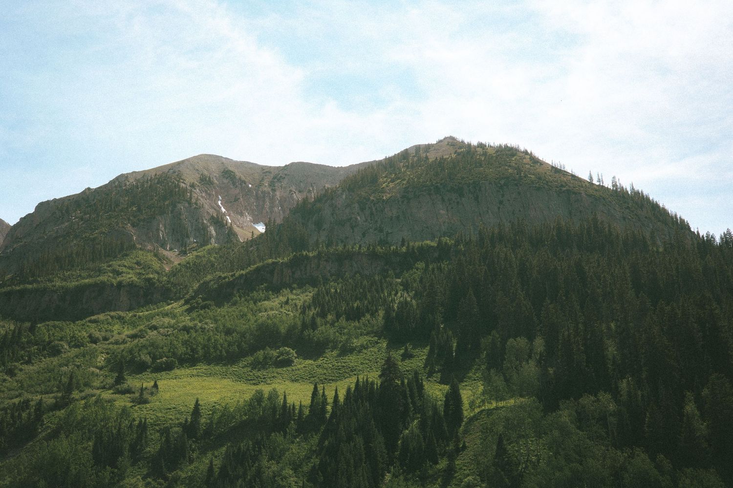 Lush green forested mountain landscape with dense pine trees and rocky peaks under a partly cloudy sky.