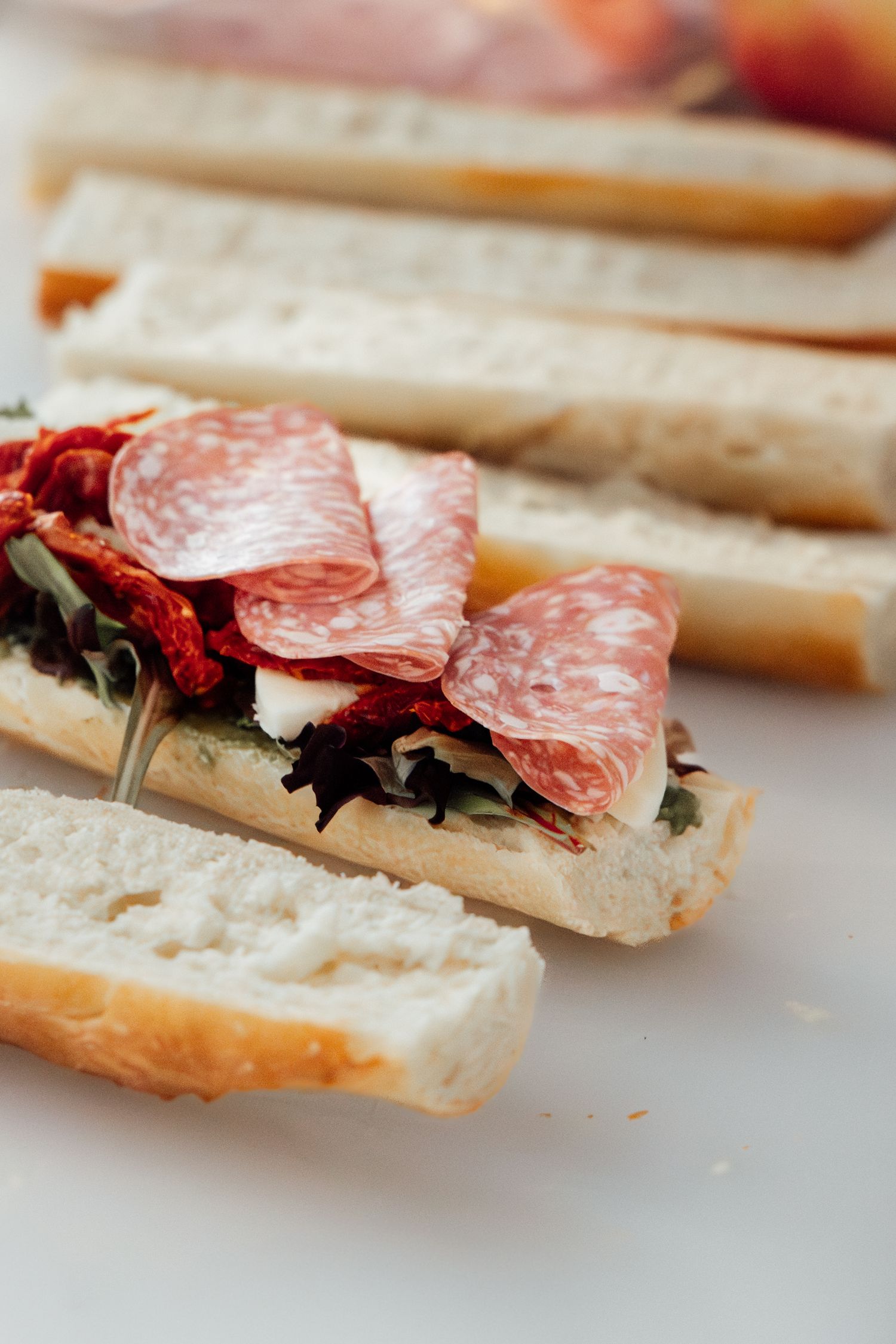 Open-faced baguette sandwich topped with salami slices, sun-dried tomatoes, and fresh greens on a white surface.