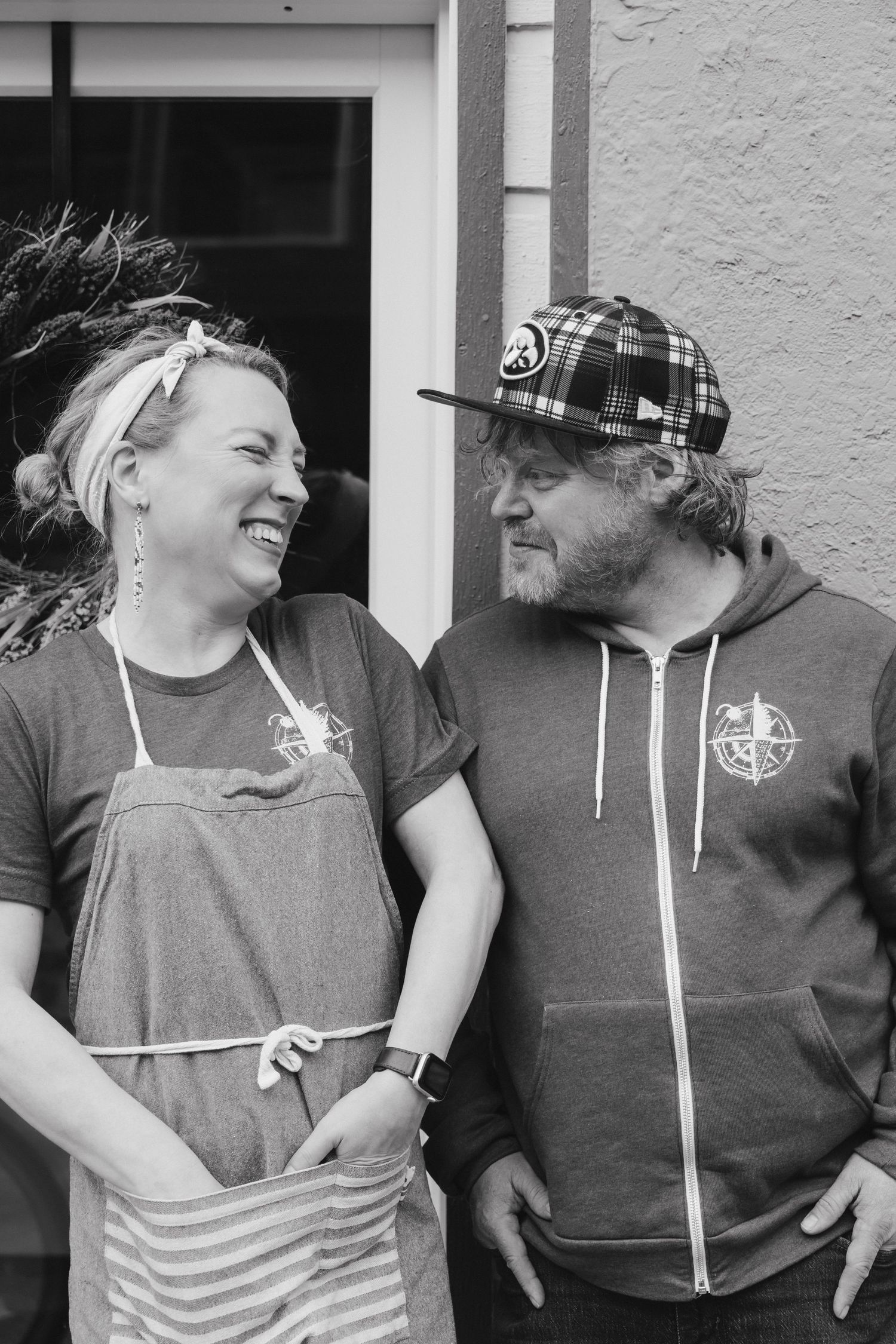 A smiling man and woman in matching branded aprons and plaid cap pose together outside a restaurant.
