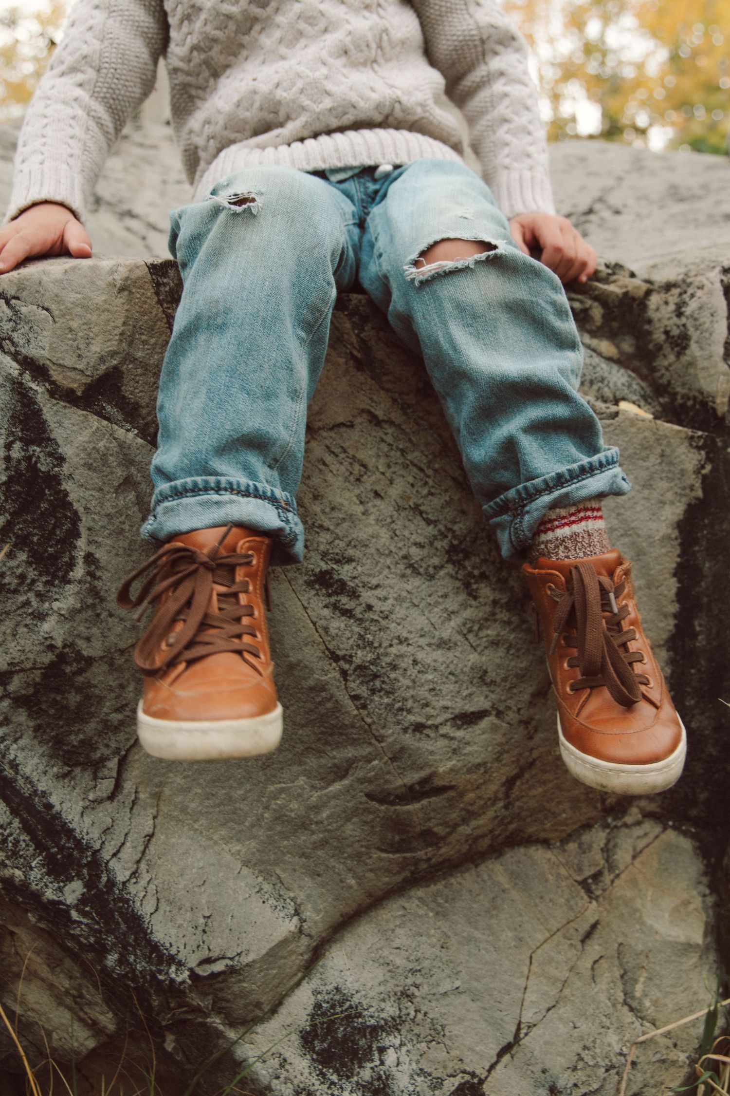 Person wearing ripped jeans and brown leather boots sits on rocky outcrop with autumn foliage in background.