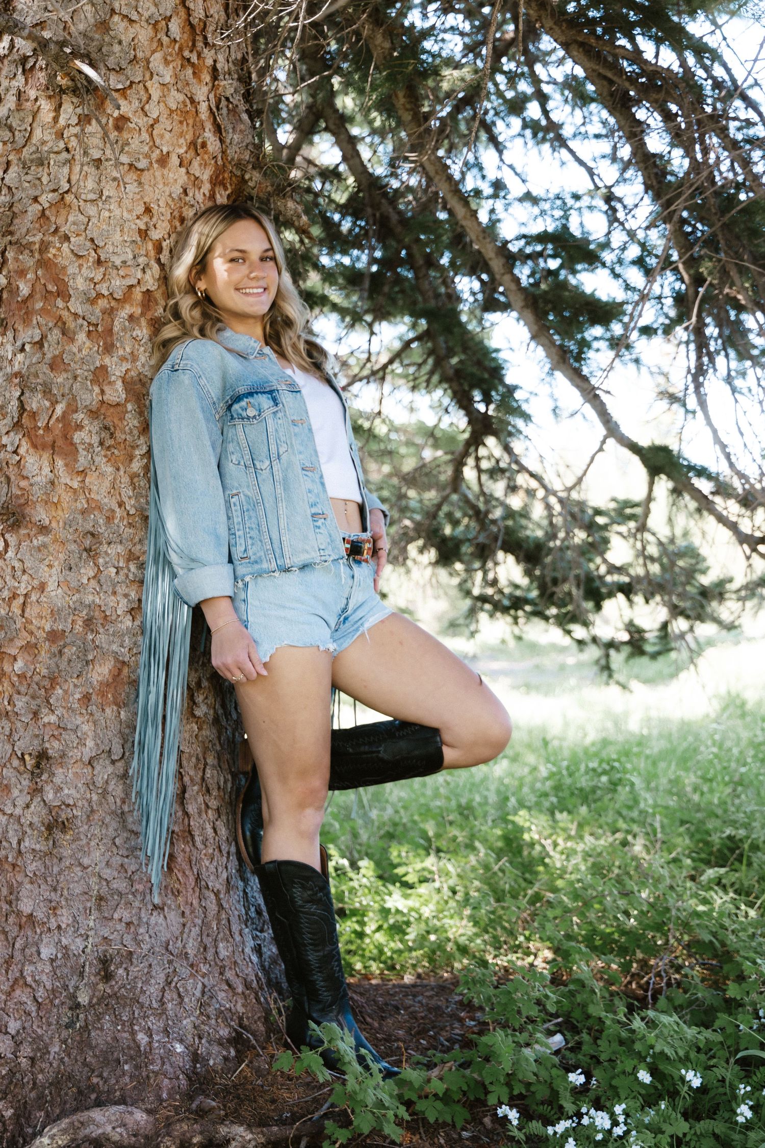 Someone in denim shorts and jacket poses casually against a large tree trunk.