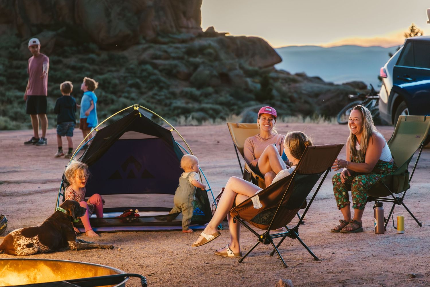 Group enjoying a camping trip at sunset with camping chairs and tent set up near mountains.