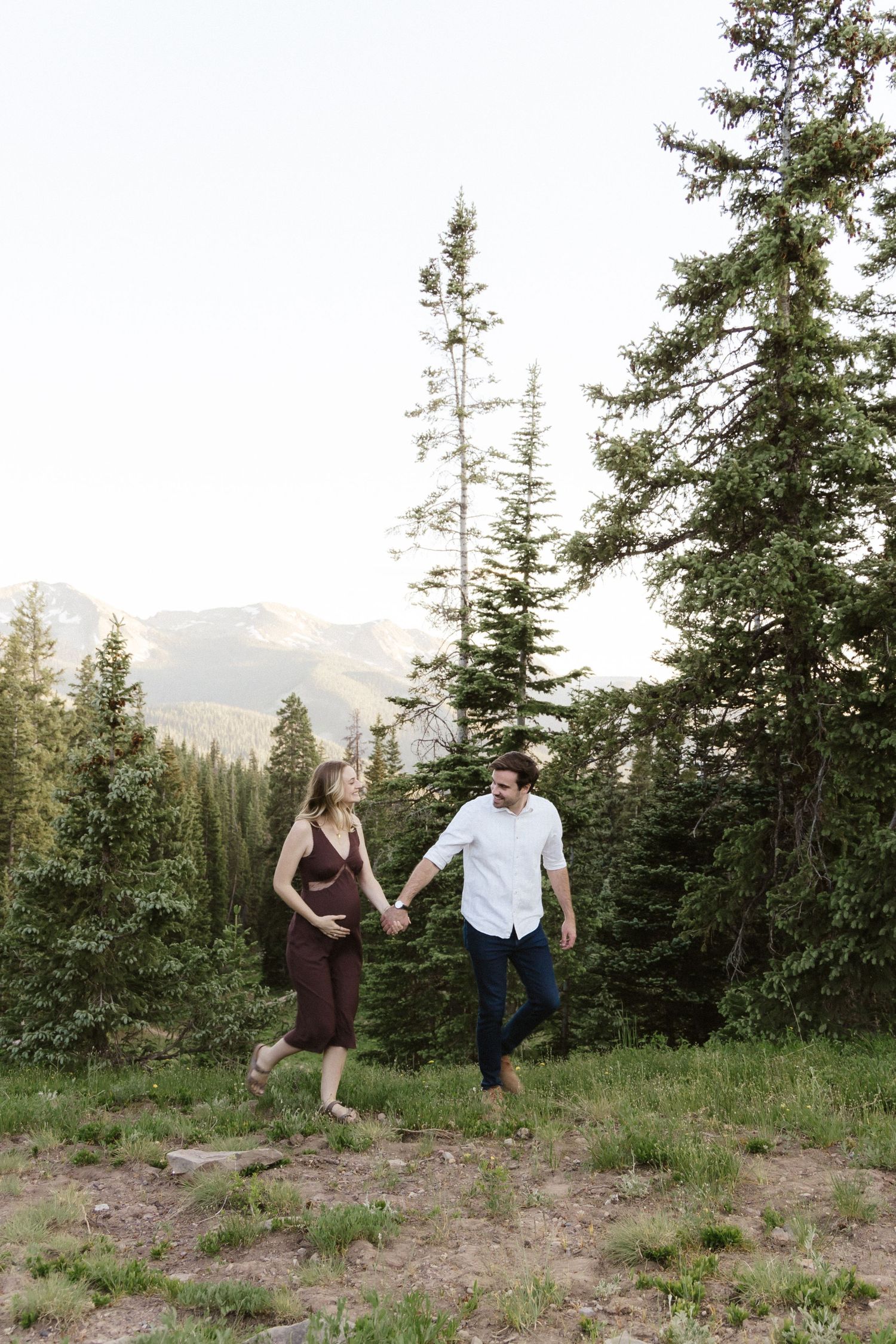 Couple walking through an alpine meadow with evergreen trees and mountain views.