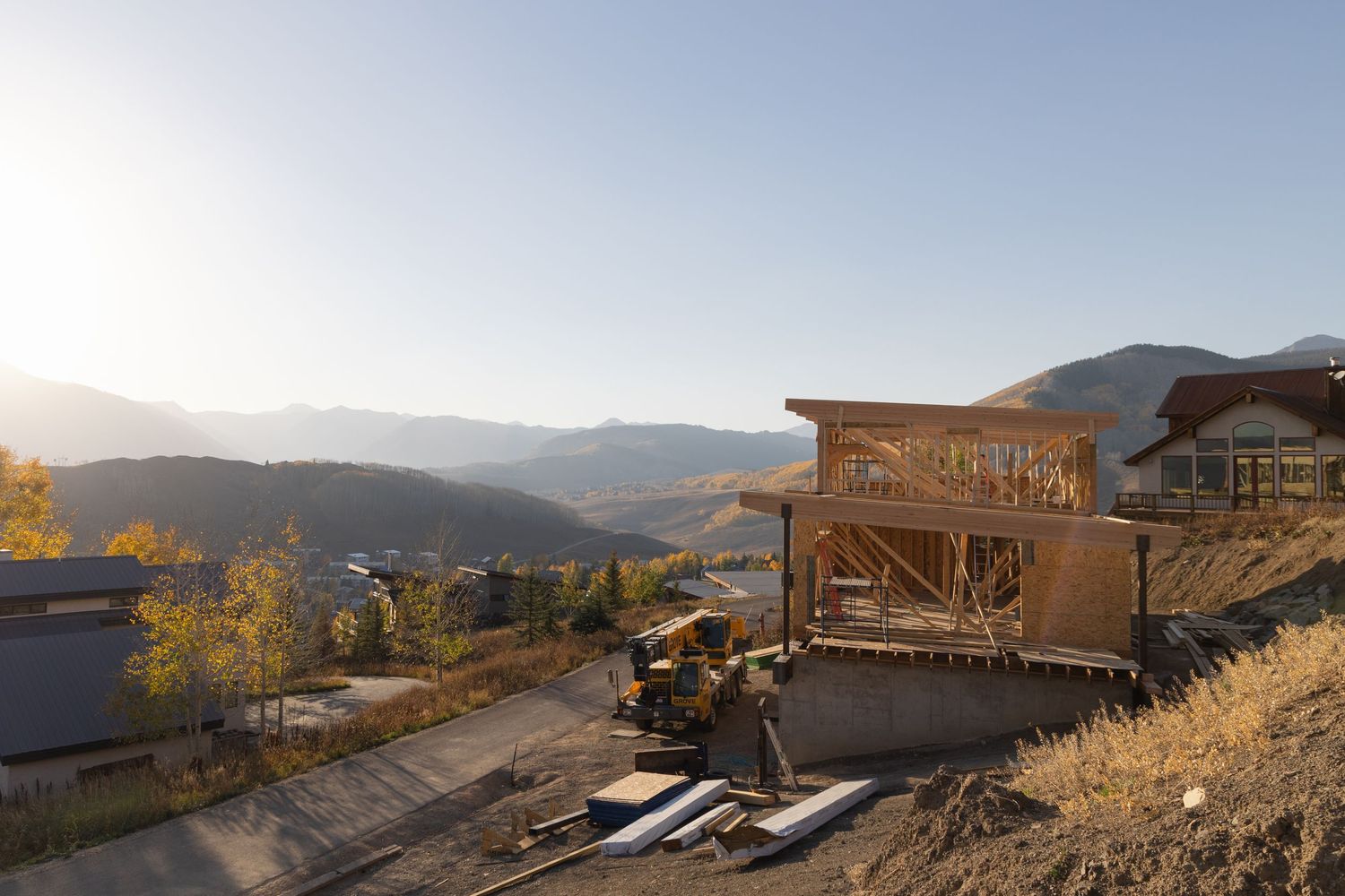Mountain home under construction with wooden framing, surrounded by fall foliage and scenic valley views at sunset.