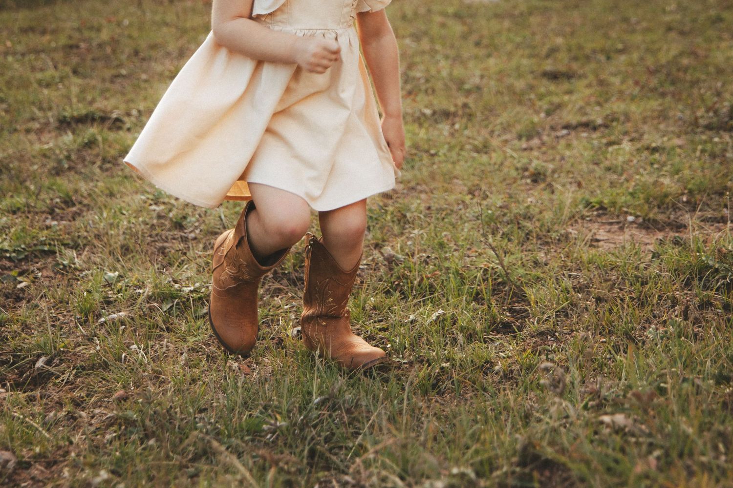 Young girl in cream dress and brown cowboy boots standing on green grass field.