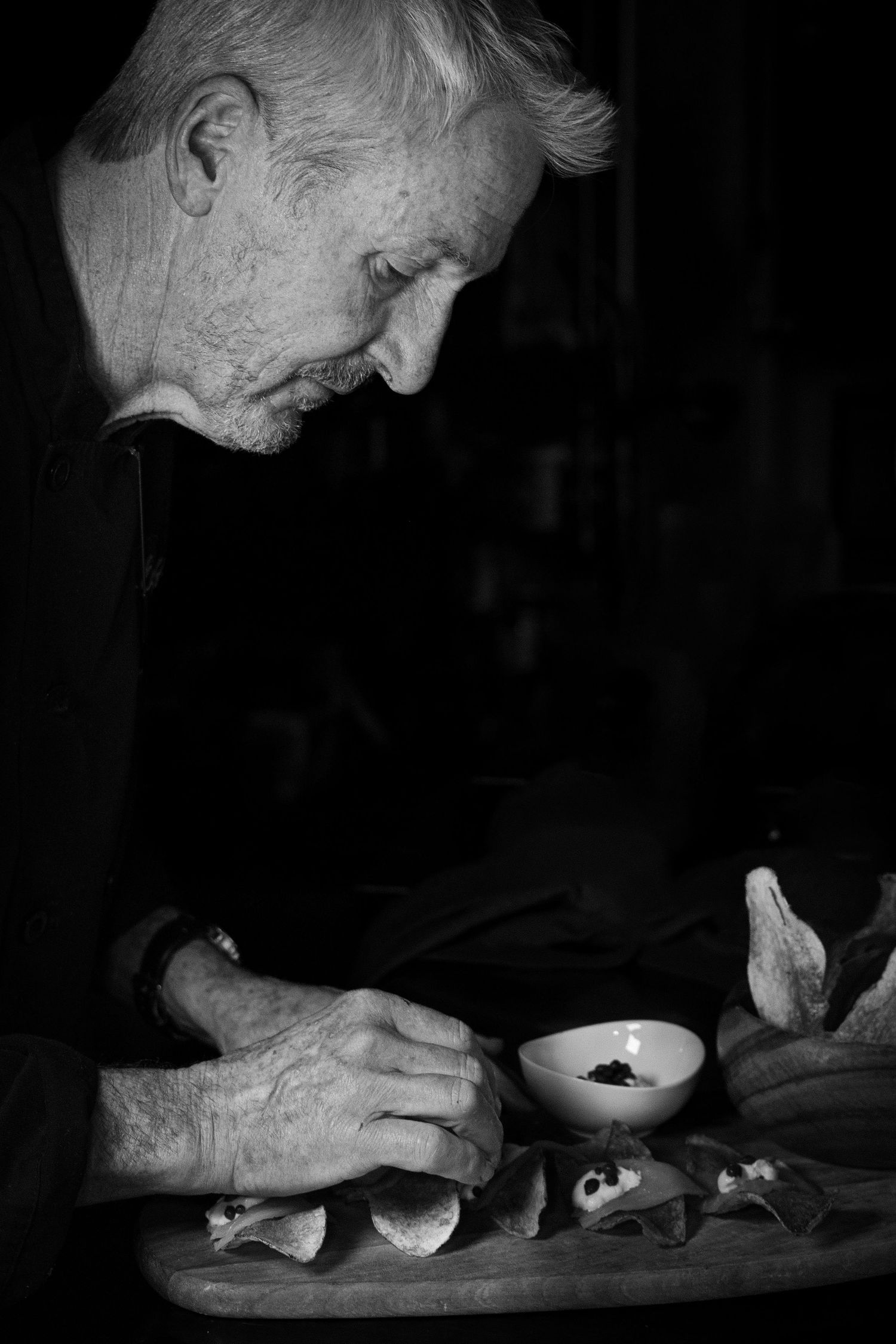 A black and white photograph of an elderly person preparing food at a wooden cutting board in dim lighting.
