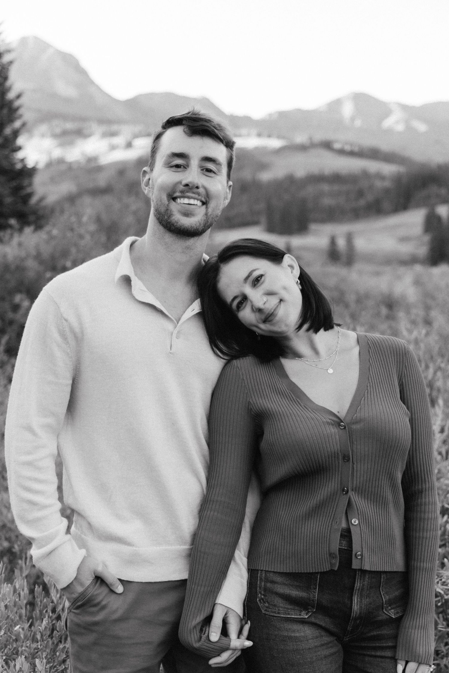 A couple poses together in a field with mountains in the background wearing coordinating white and brown outfits.