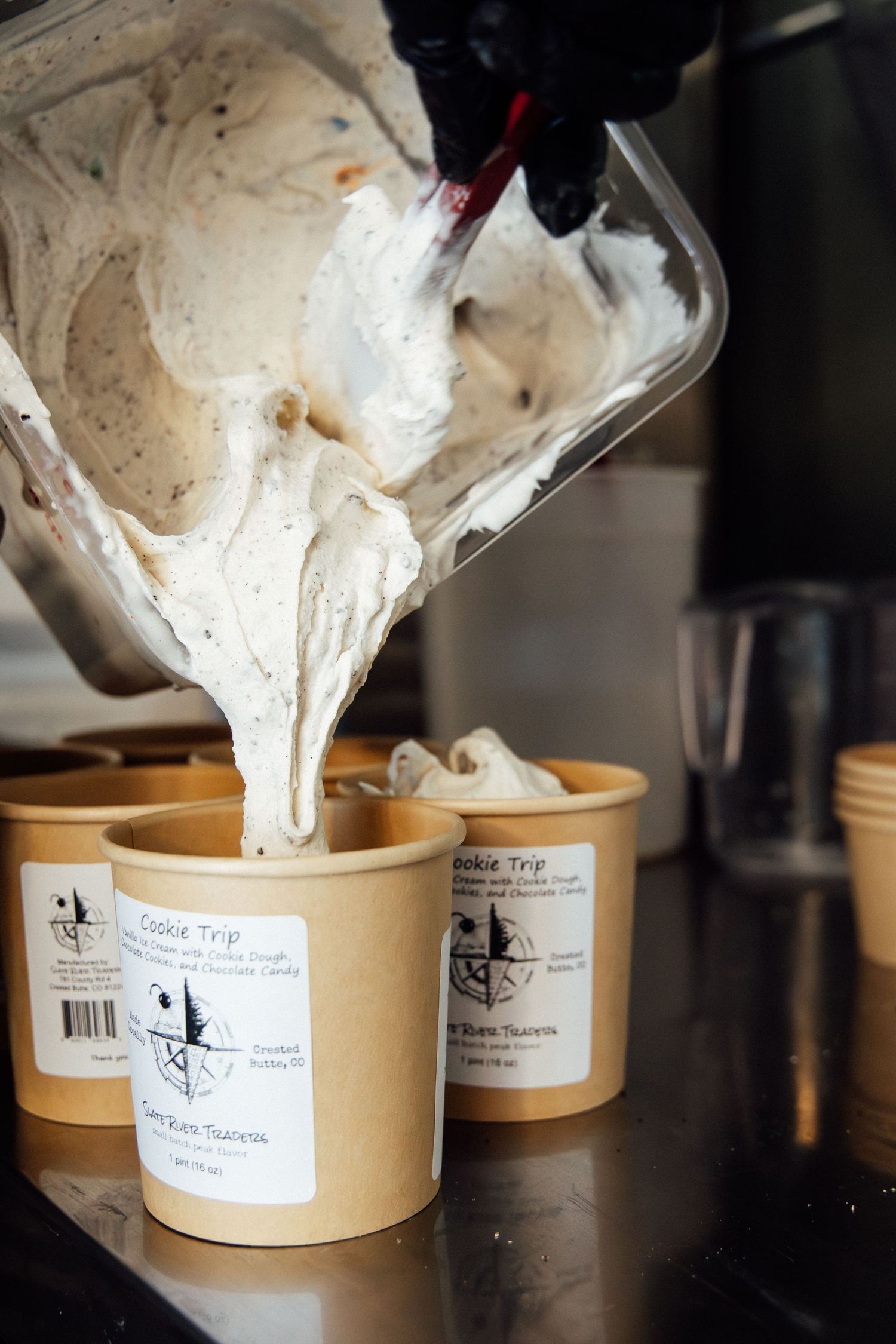 Gloved hands pouring creamy Cookie Trip ice cream into kraft paper cups on a stainless steel counter.