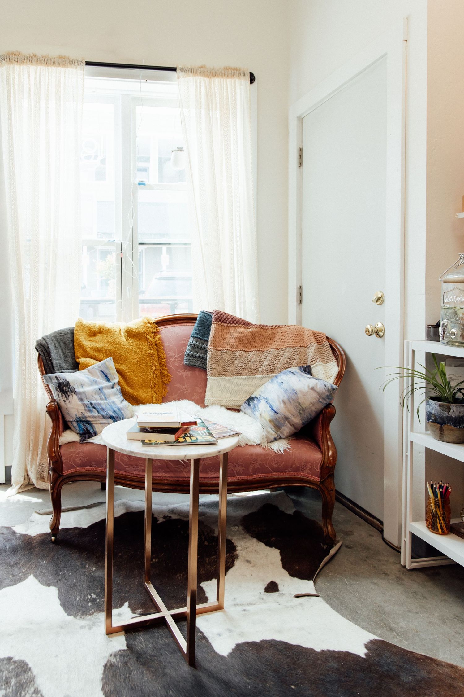 Bohemian living room corner with vintage loveseat, colorful pillows, cowhide rug, and gold side table near sheer curtains.