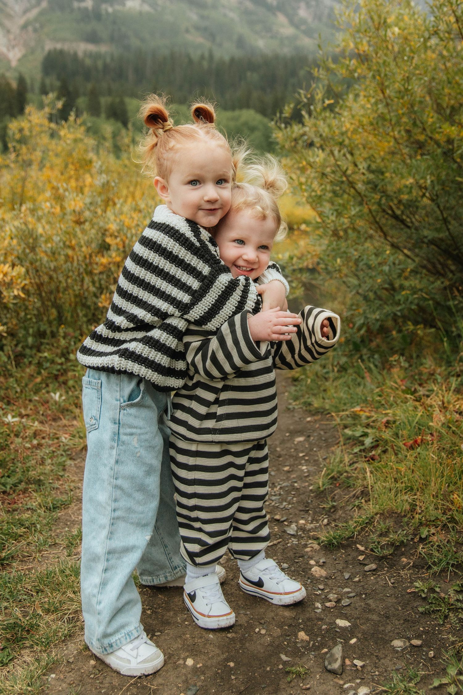 Two small children in striped clothing hugging outdoors with mountain backdrop.