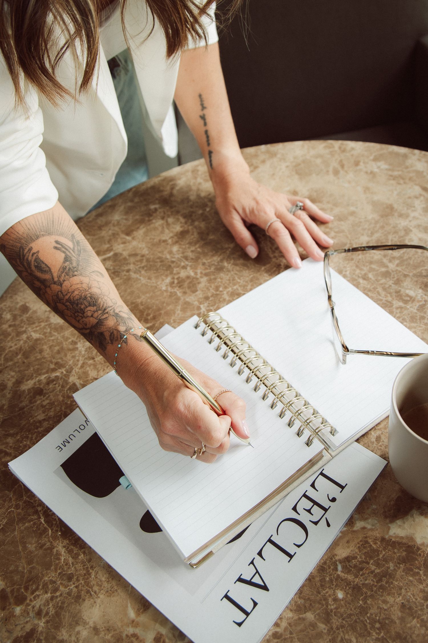 Tattooed woman writing in spiral notebook at marble table with coffee cup and L'Eclat magazine.