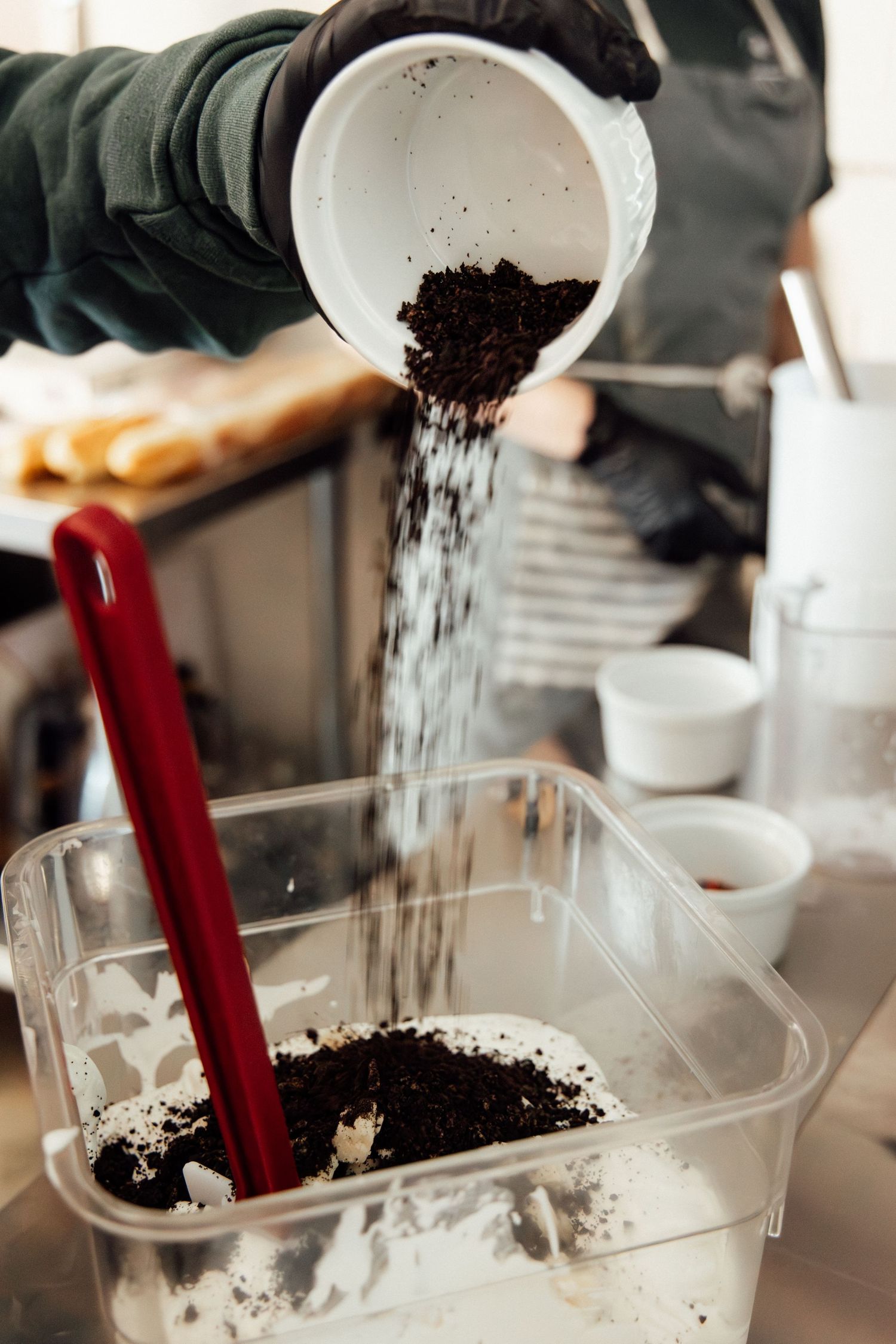 A chef in black gloves pours dark coffee grounds into a clear container with a red spatula in a professional kitchen.