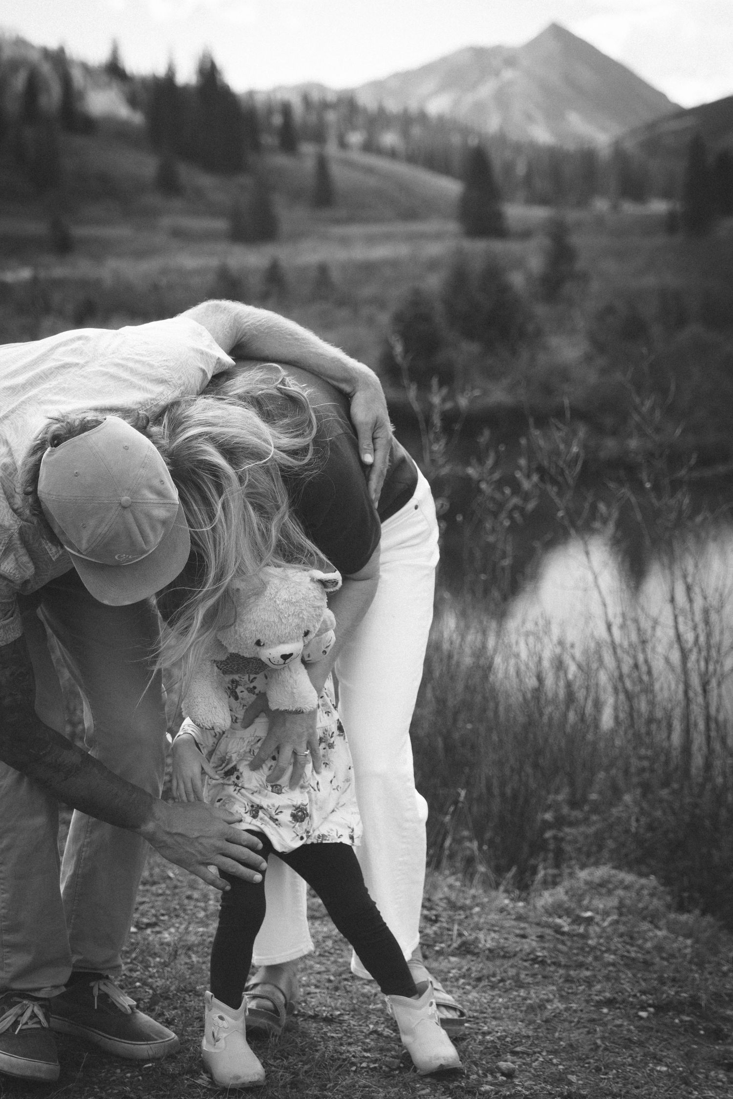Black and white photo of family with young child holding teddy bear near mountain lake surrounded by pine trees.