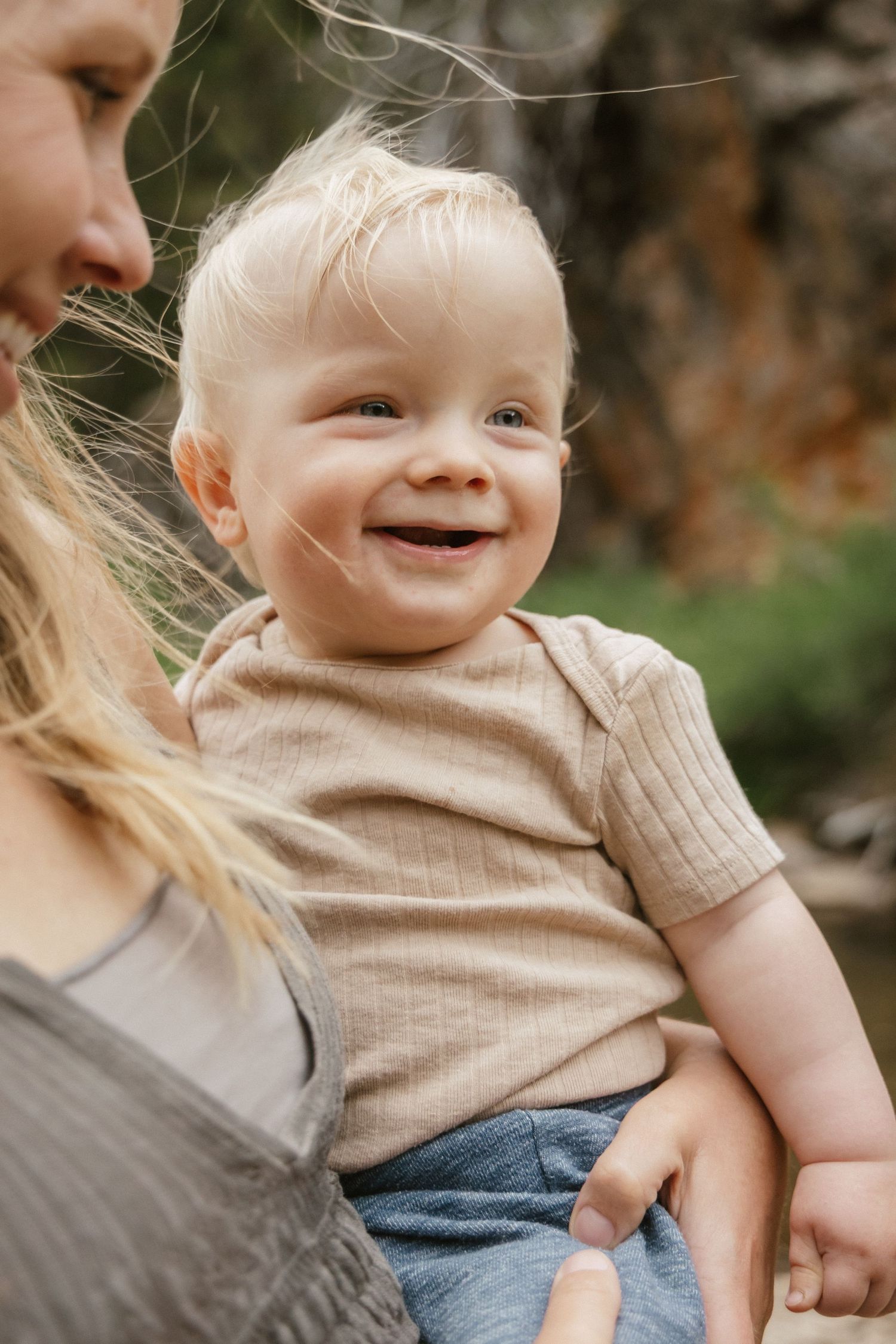 A close-up portrait of a smiling toddler in a beige shirt being held outdoors.