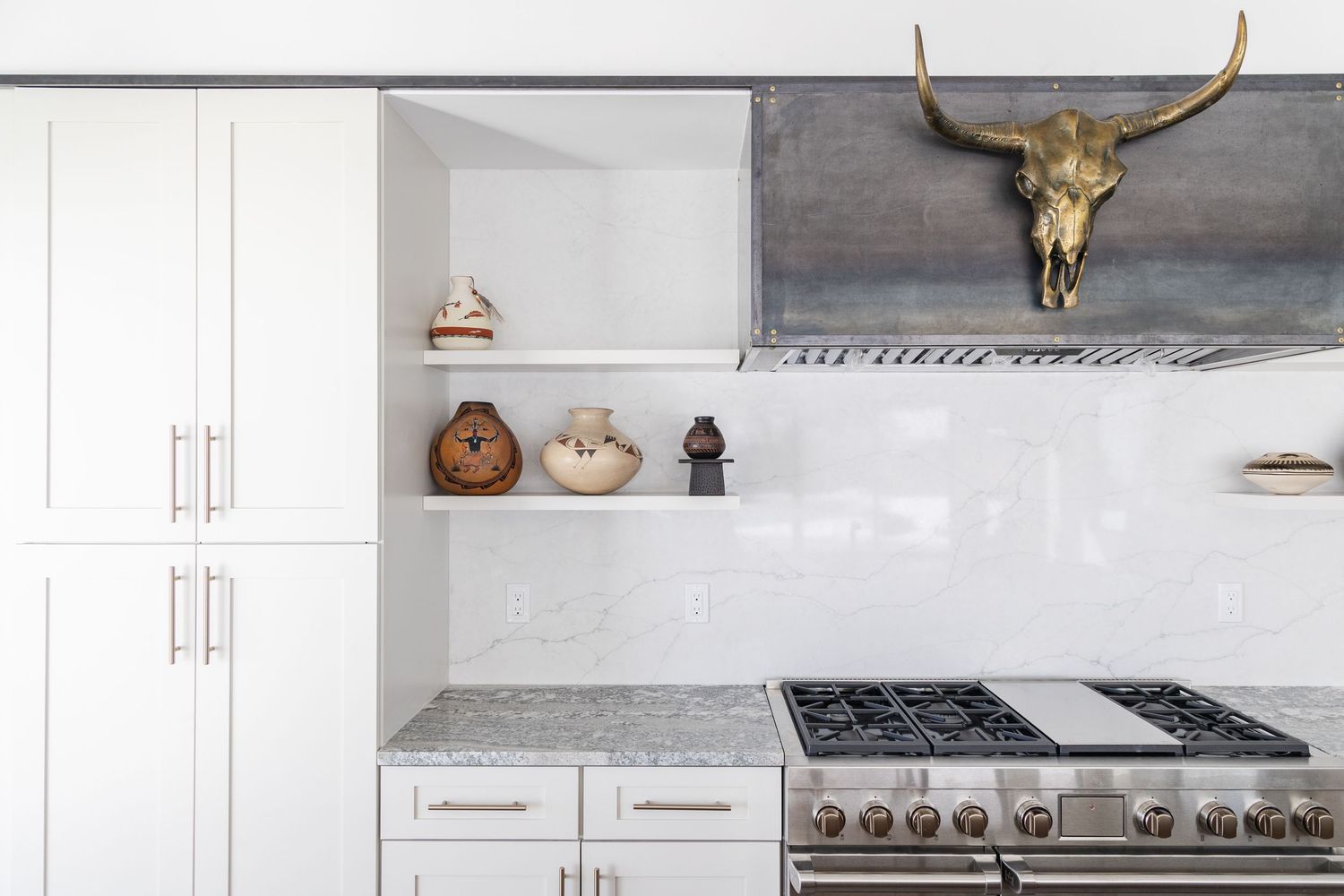 White kitchen with open shelves displaying pottery, marble countertops, stainless steel range, and a decorative bull skull above the hood.