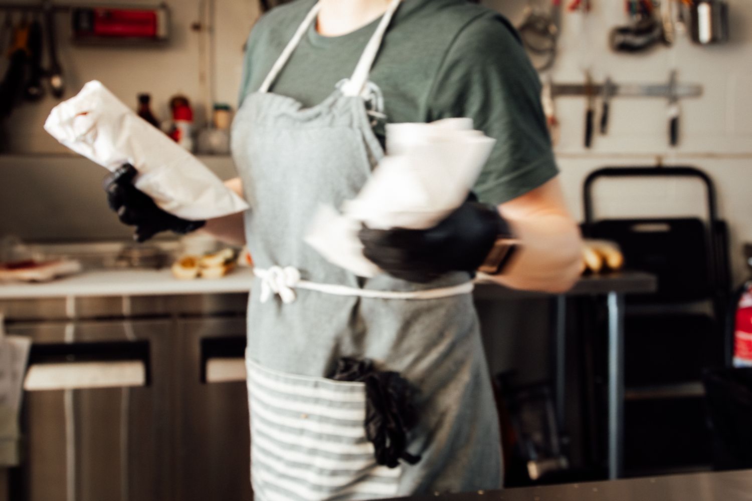 A cook in a gray apron and black gloves holds white kitchen towels in a professional kitchen.