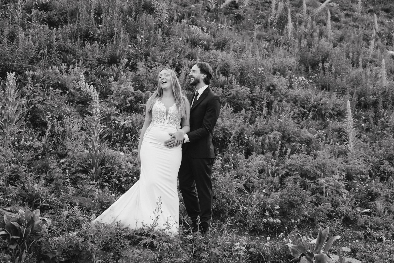 Black and white photo of a bride and groom standing together in a lush green hillside, sharing a romantic moment.