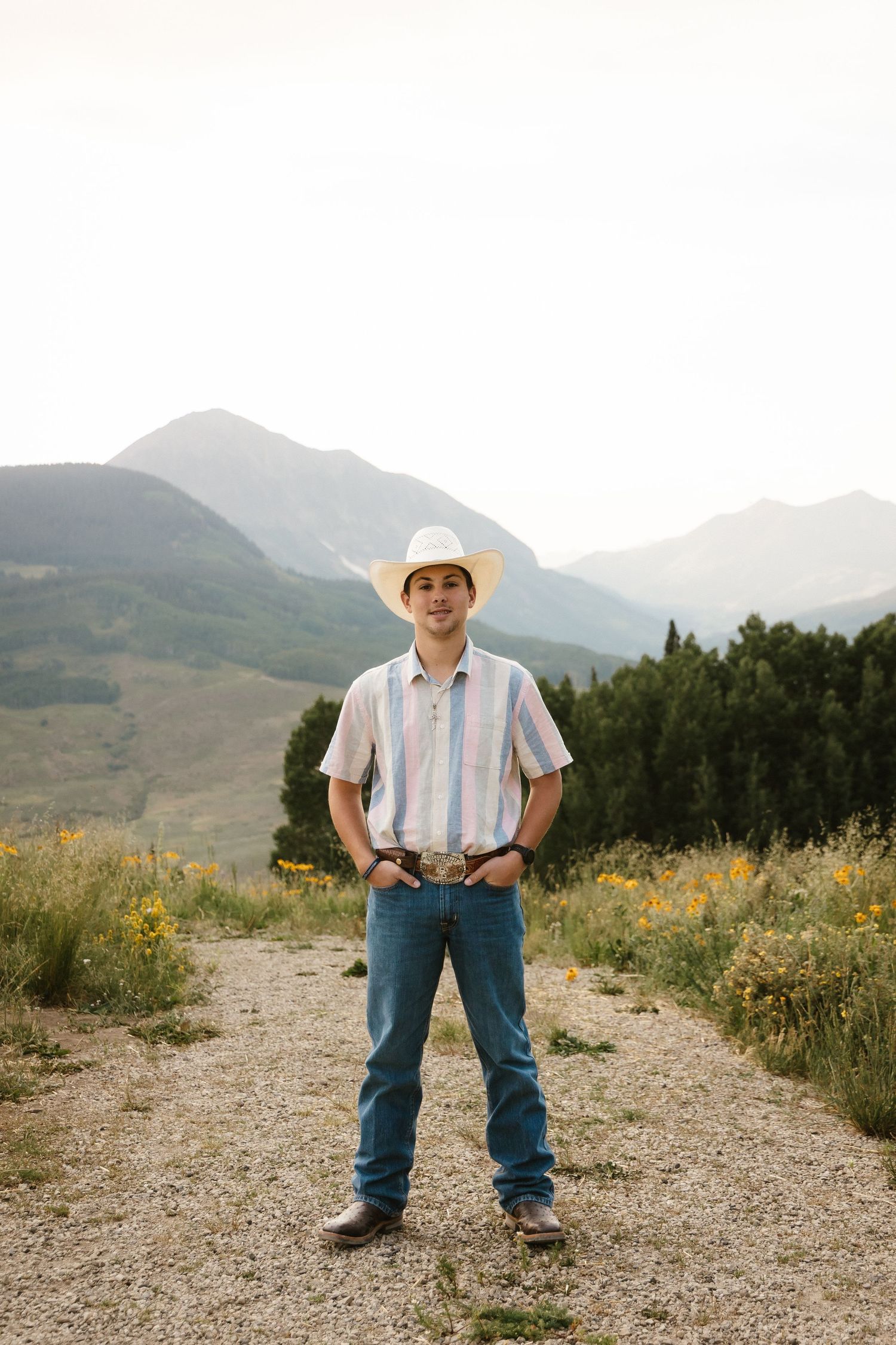 A young cowboy stands confidently against a mountainous backdrop on a gravel path.