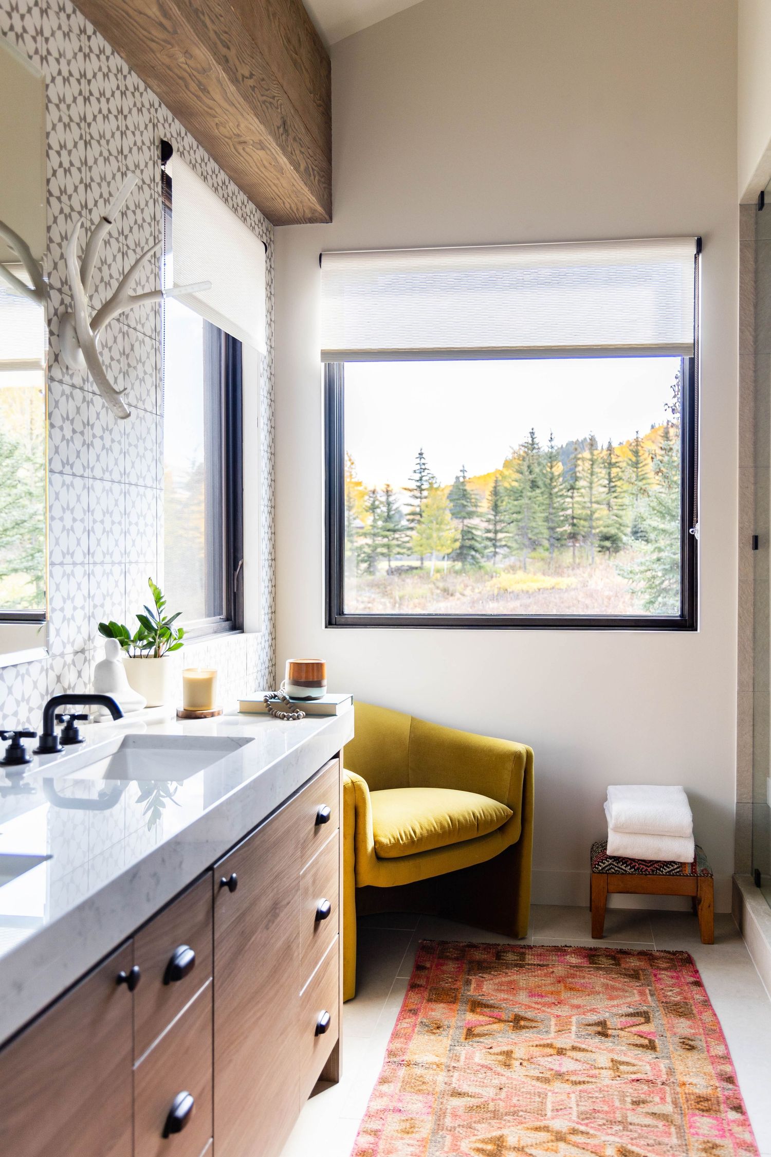 Modern bathroom with wood vanity, yellow accent chair, colorful rug, and scenic forest view through black-framed window.