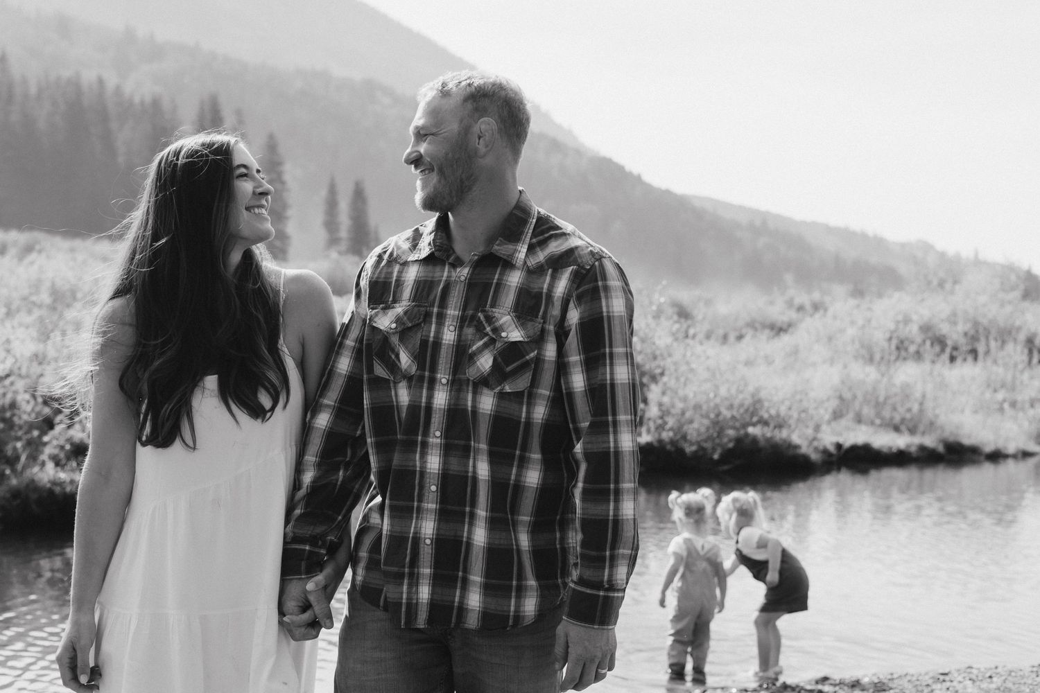 A couple shares a tender moment by a lake while children play in the background.