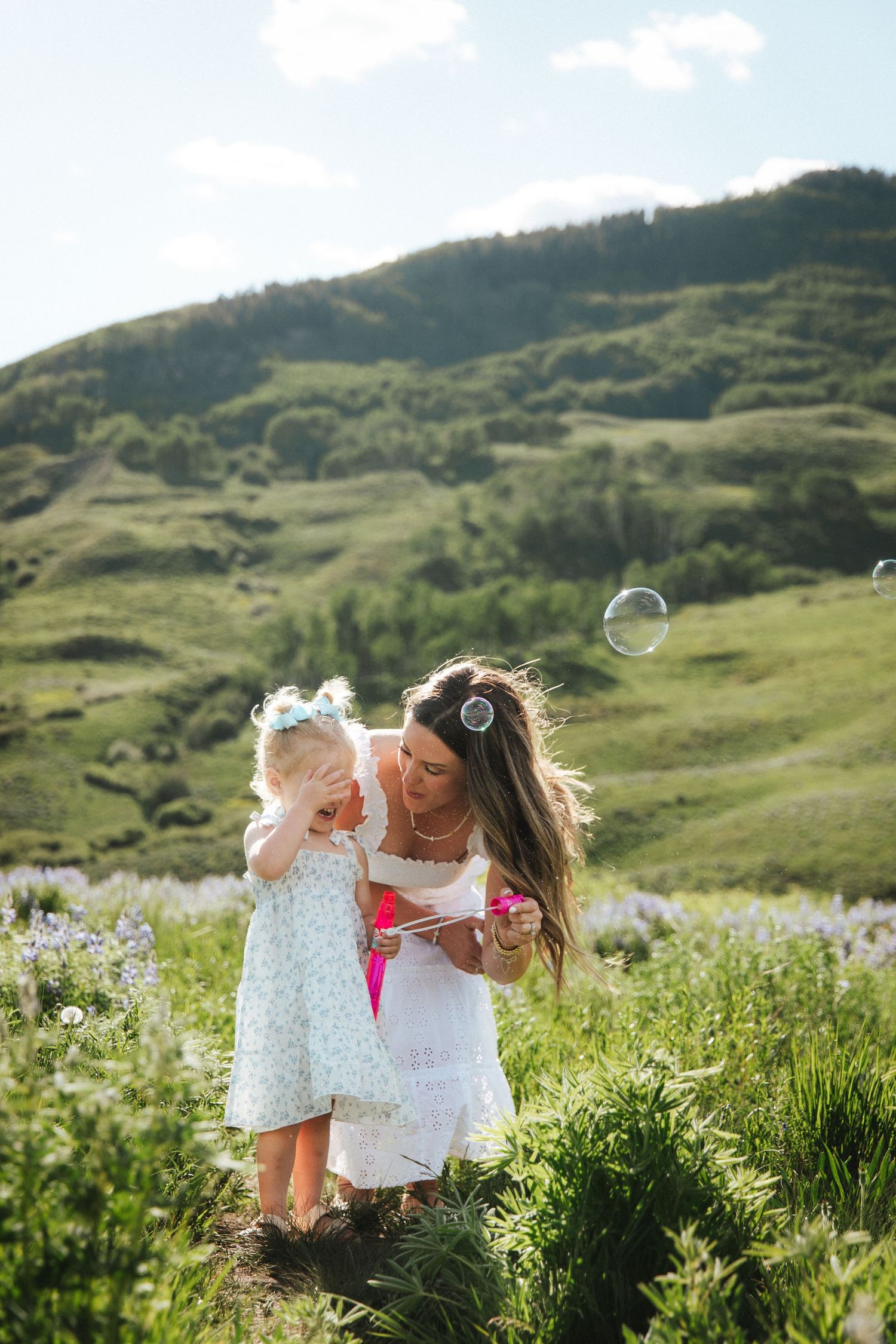 Mother and daughter blowing soap bubbles in a green meadow with rolling hills on a sunny day.