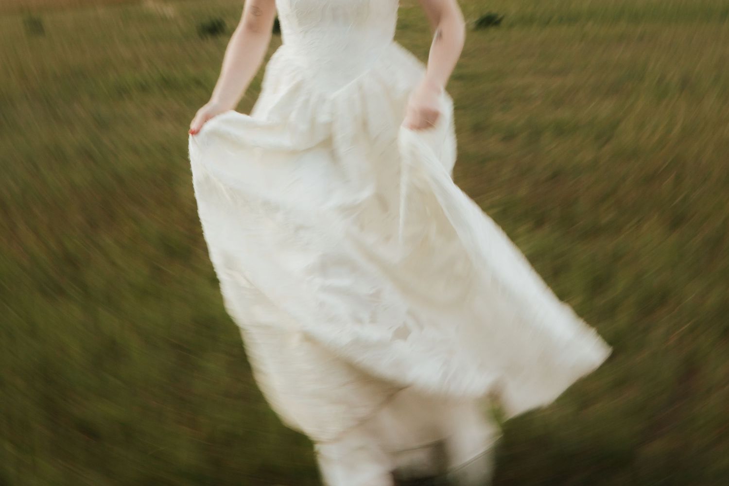 A bride in a flowing white wedding dress runs through a lush green field, captured in a dreamy motion blur.