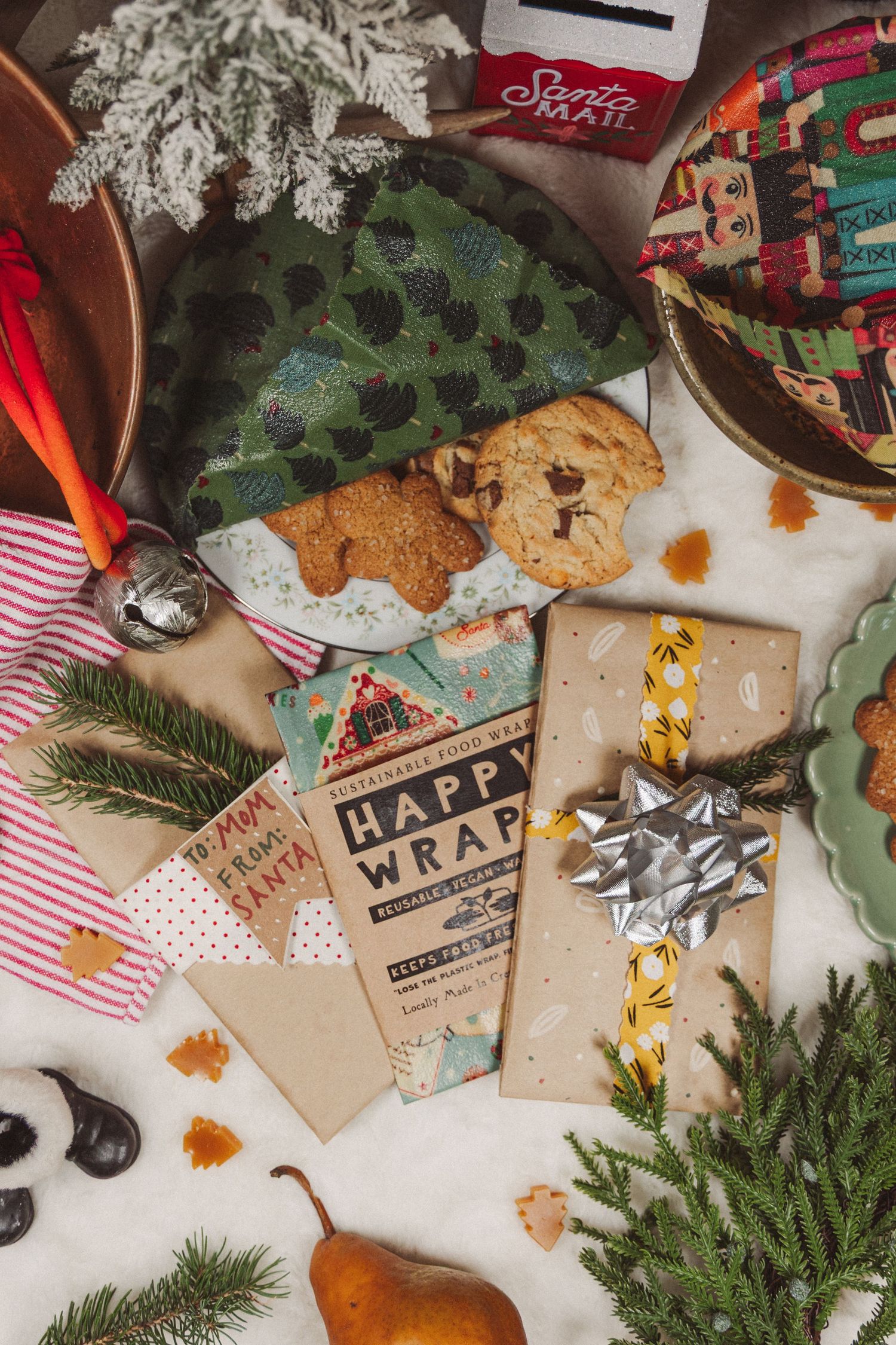 Festive holiday scene with Christmas cookies, evergreen branches, lights and gift wrapping supplies on a rustic surface.